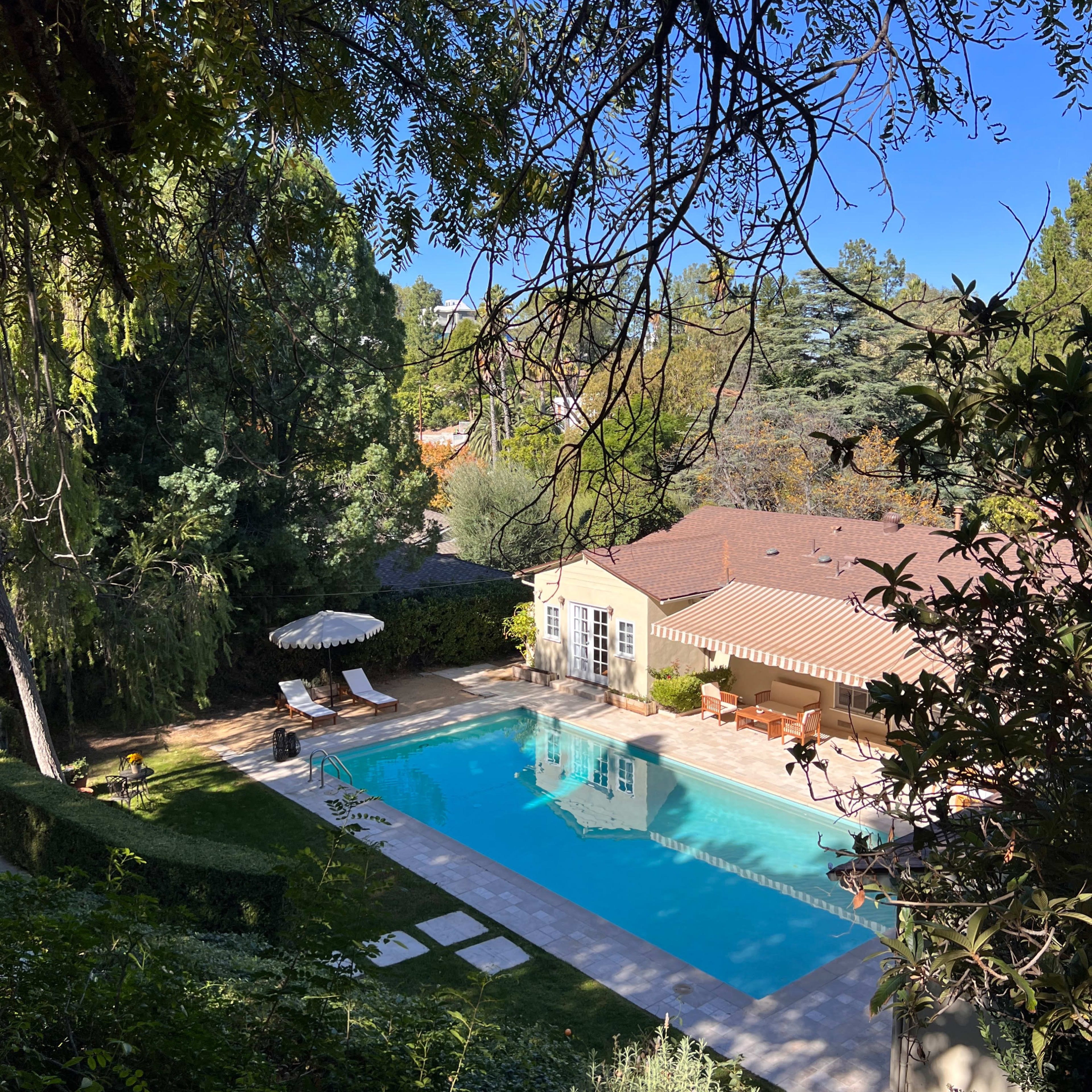 A clear swimming pool is situated beside a house with a red-tiled roof, surrounded by greenery and lawn chairs.