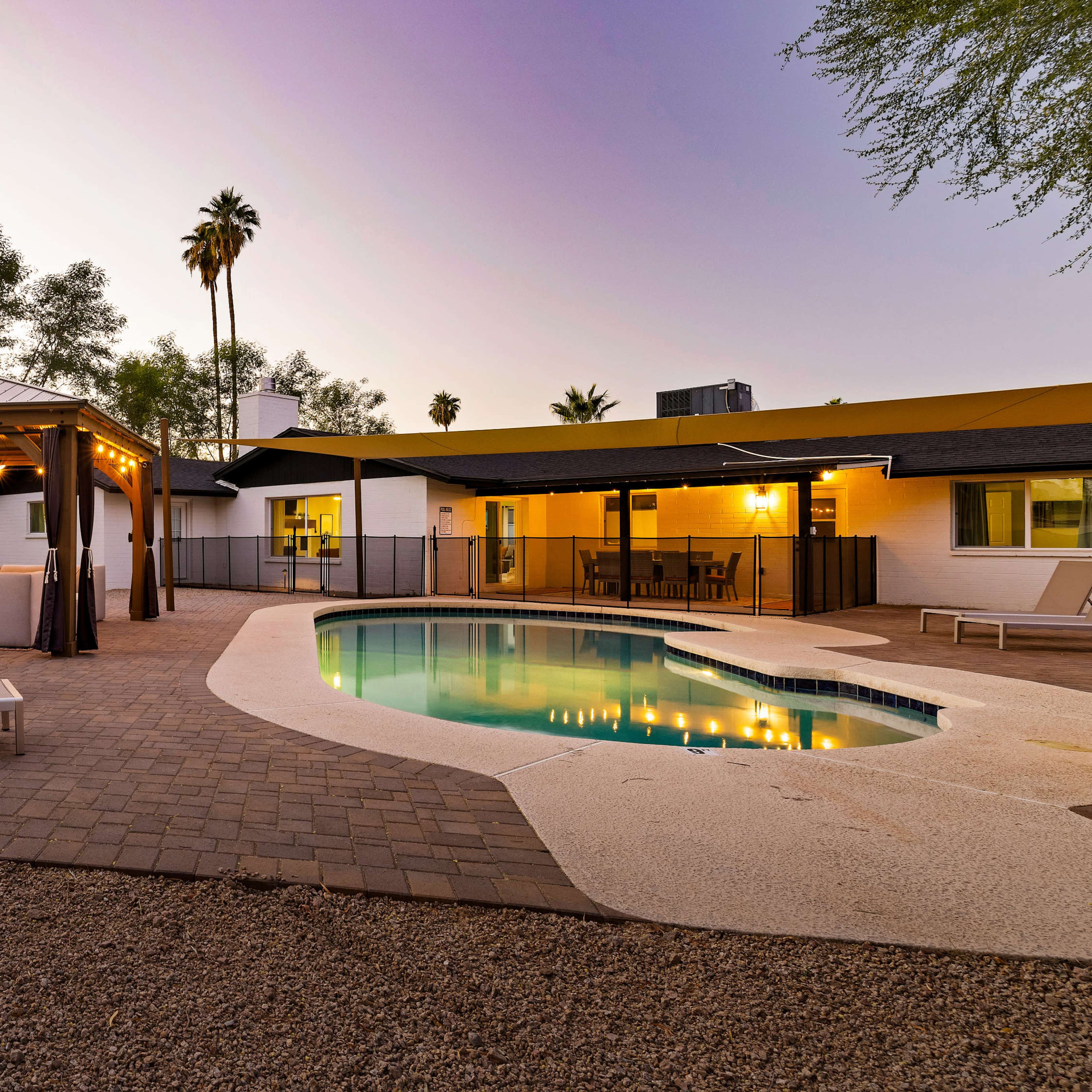 A backyard with a swimming pool, a gazebo with seating, and palm trees under a twilight sky.