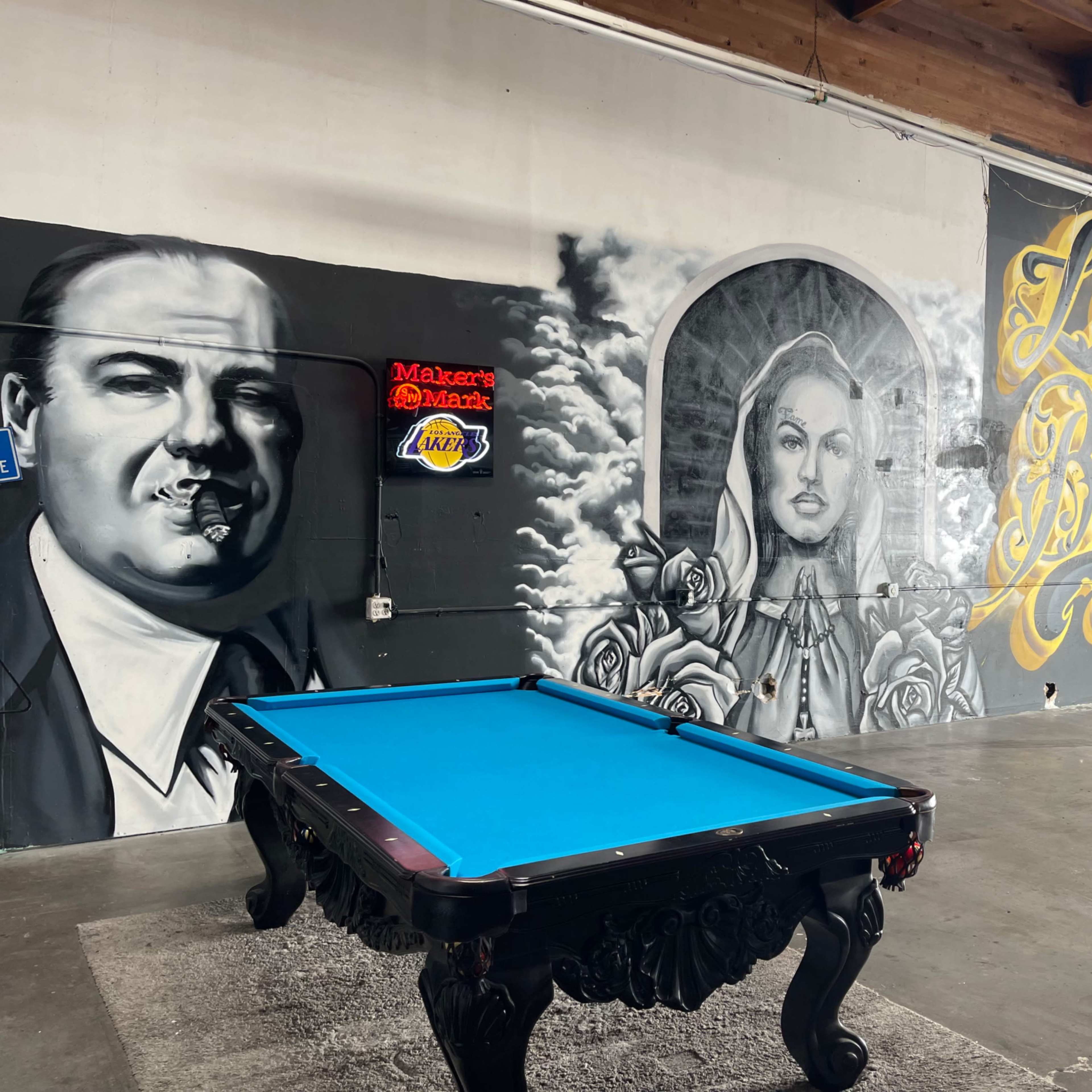 A pool table in an industrial space featuring large murals of a man with a cigar, a woman surrounded by roses, and the words "Long Beach."
