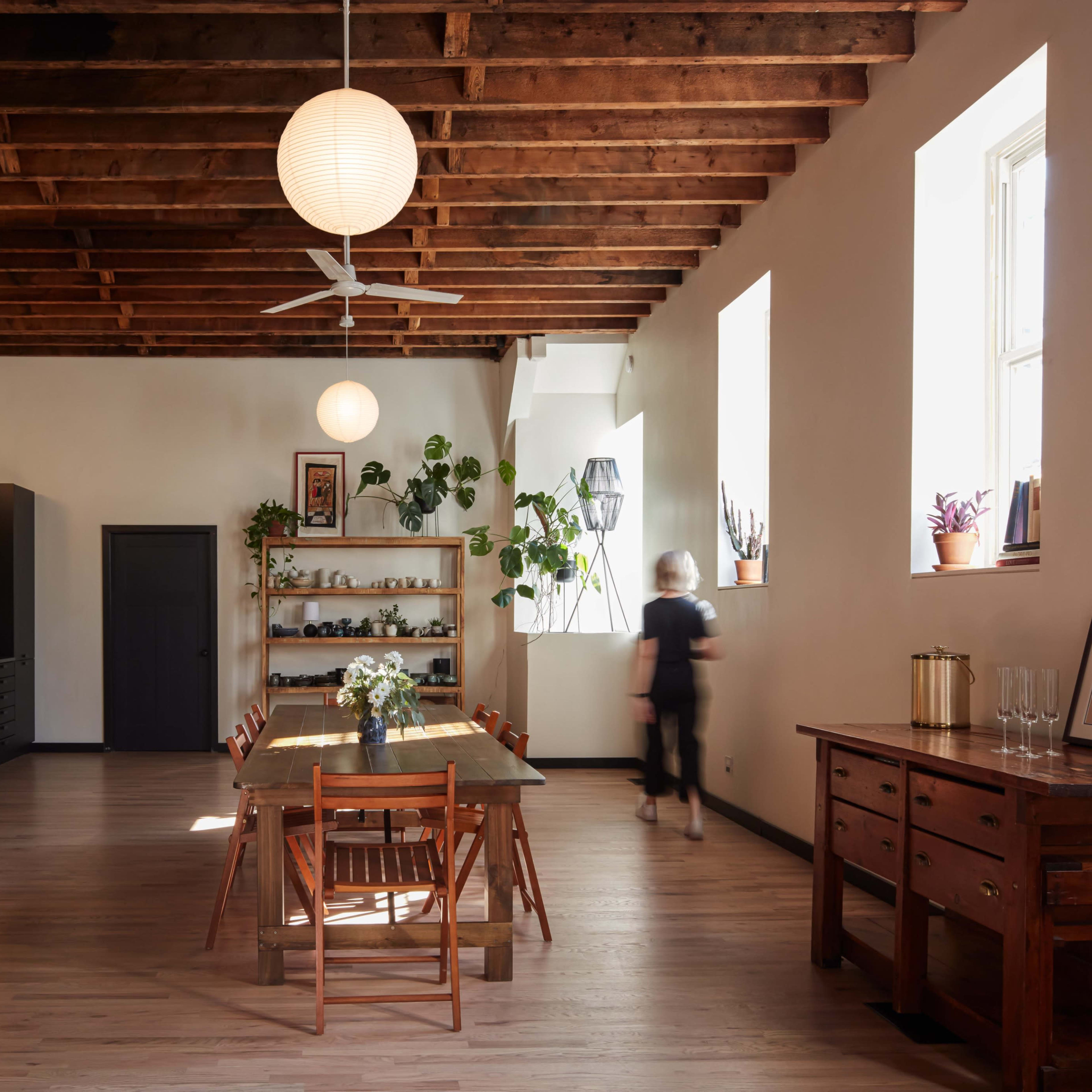 A spacious kitchen features a long wooden dining table surrounded by chairs, with open shelving, a minimalist black cabinetry setup, and large windows providing natural light.