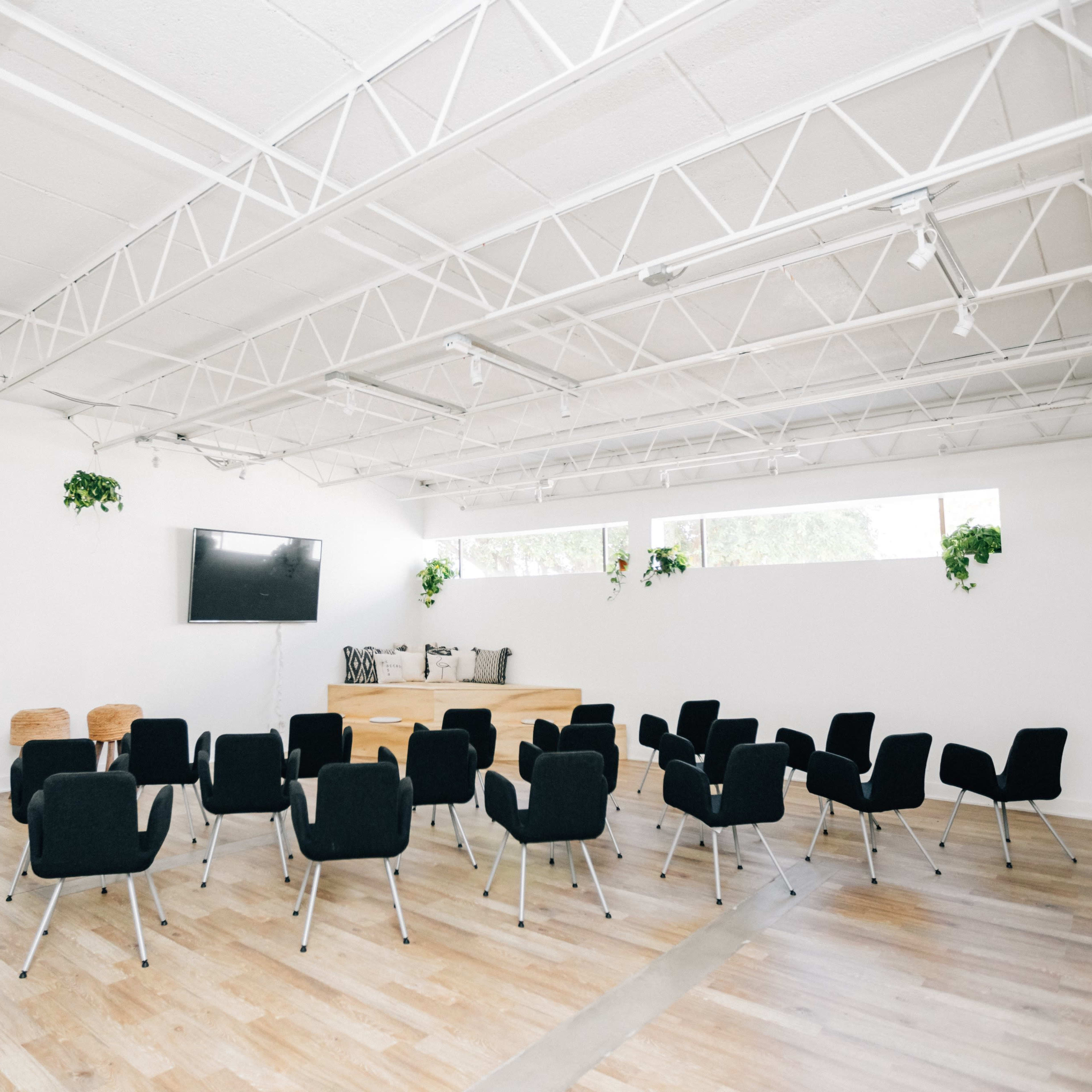 A minimalistic meeting room features a circular arrangement of black chairs facing a large screen, with potted plants on the walls and bright natural light from the windows.