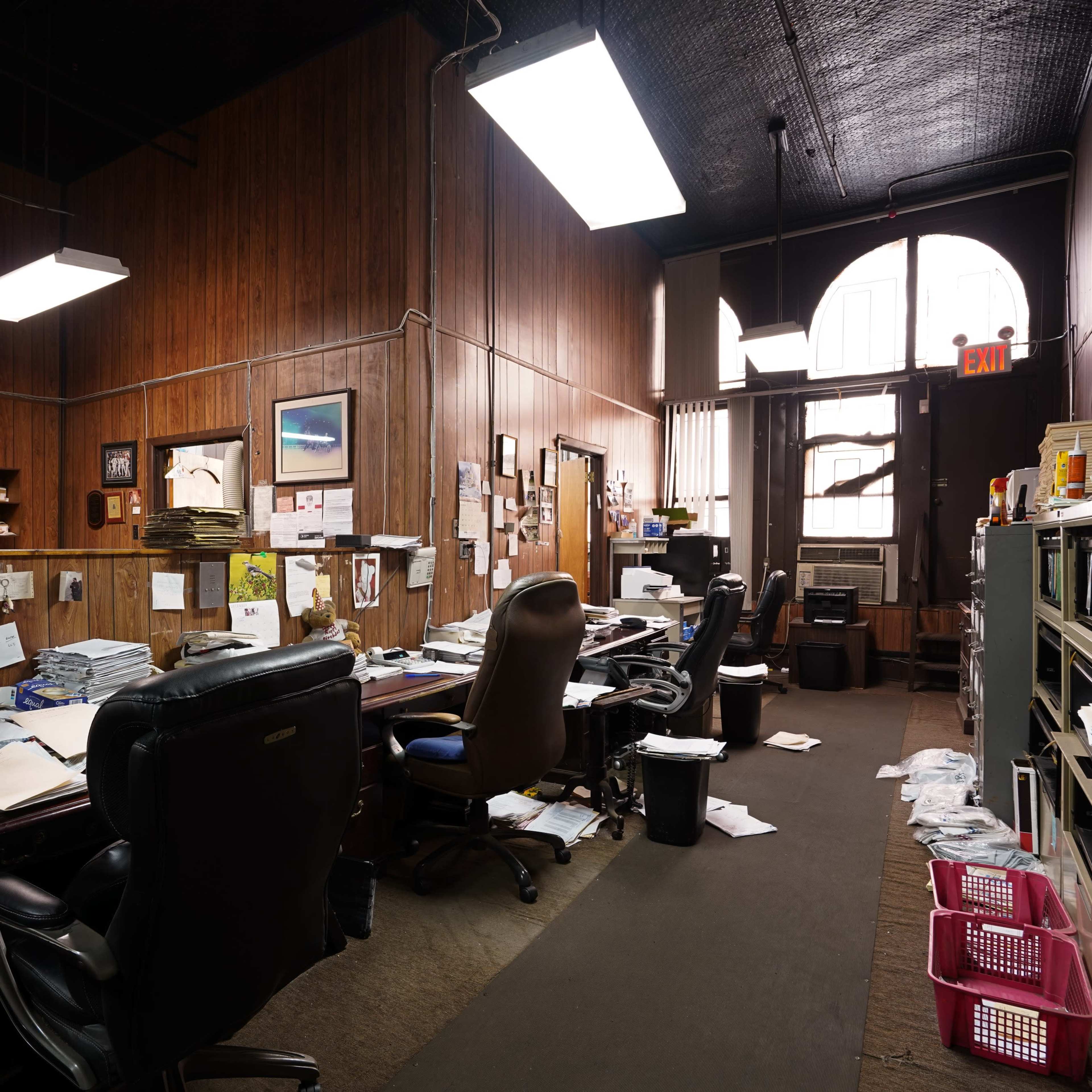 A cluttered office space with wooden paneling, multiple desks, and scattered papers throughout.