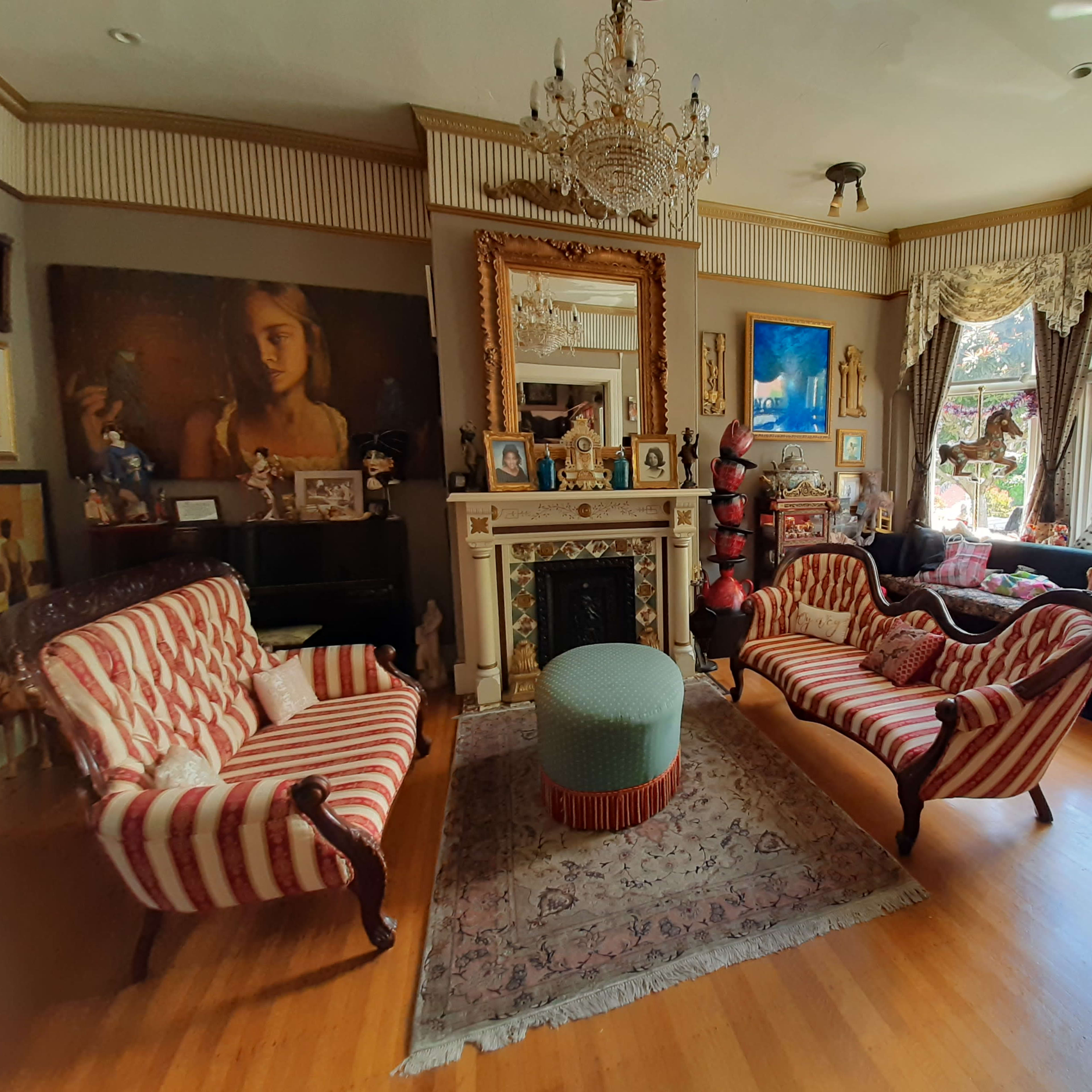 The room features a mix of striped sofas and a round ottoman, with a decorative fireplace and various art pieces adorning the walls.