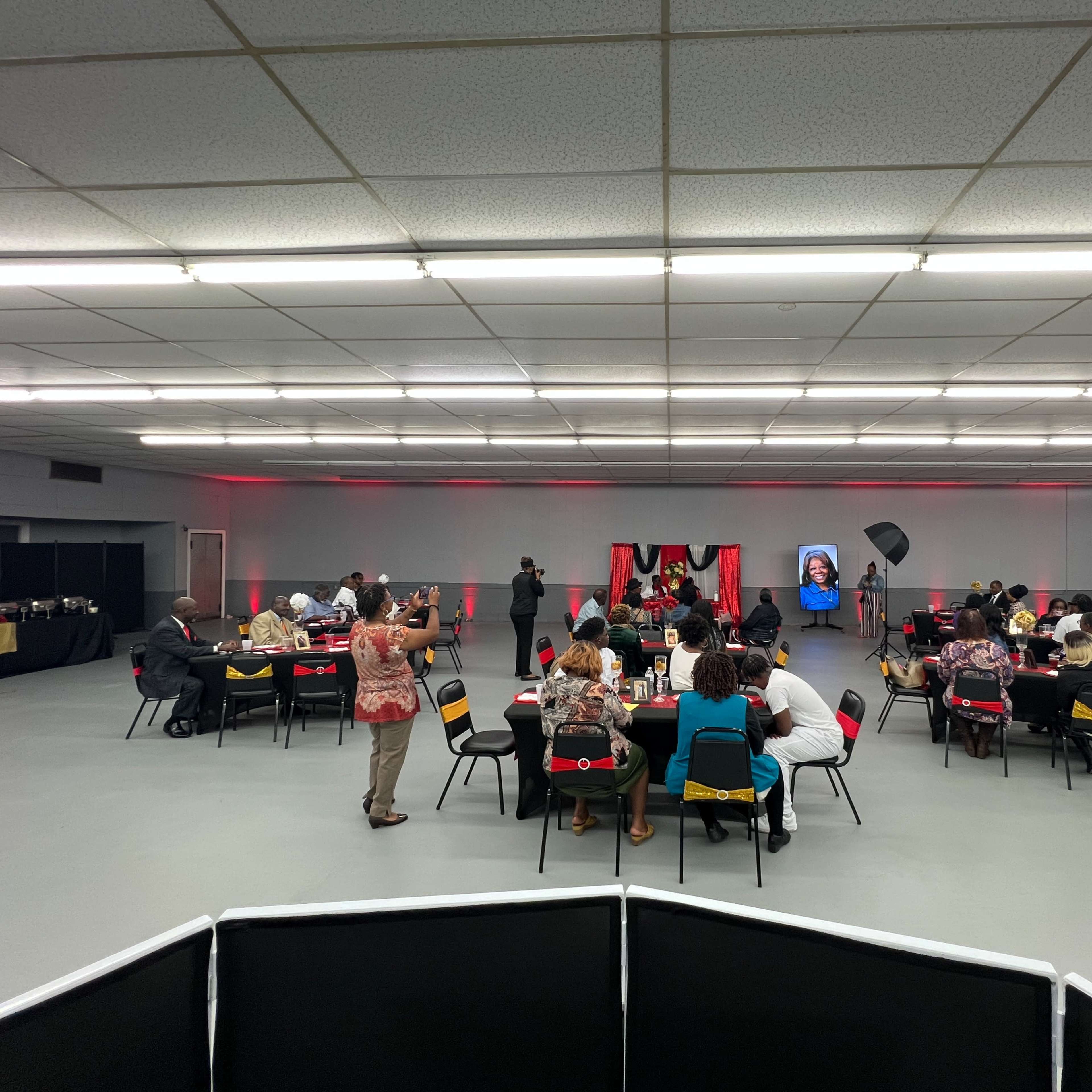 A gathering is taking place in a spacious room with tables arranged for attendees, who are engaged in various activities while a presentation occurs at the front.