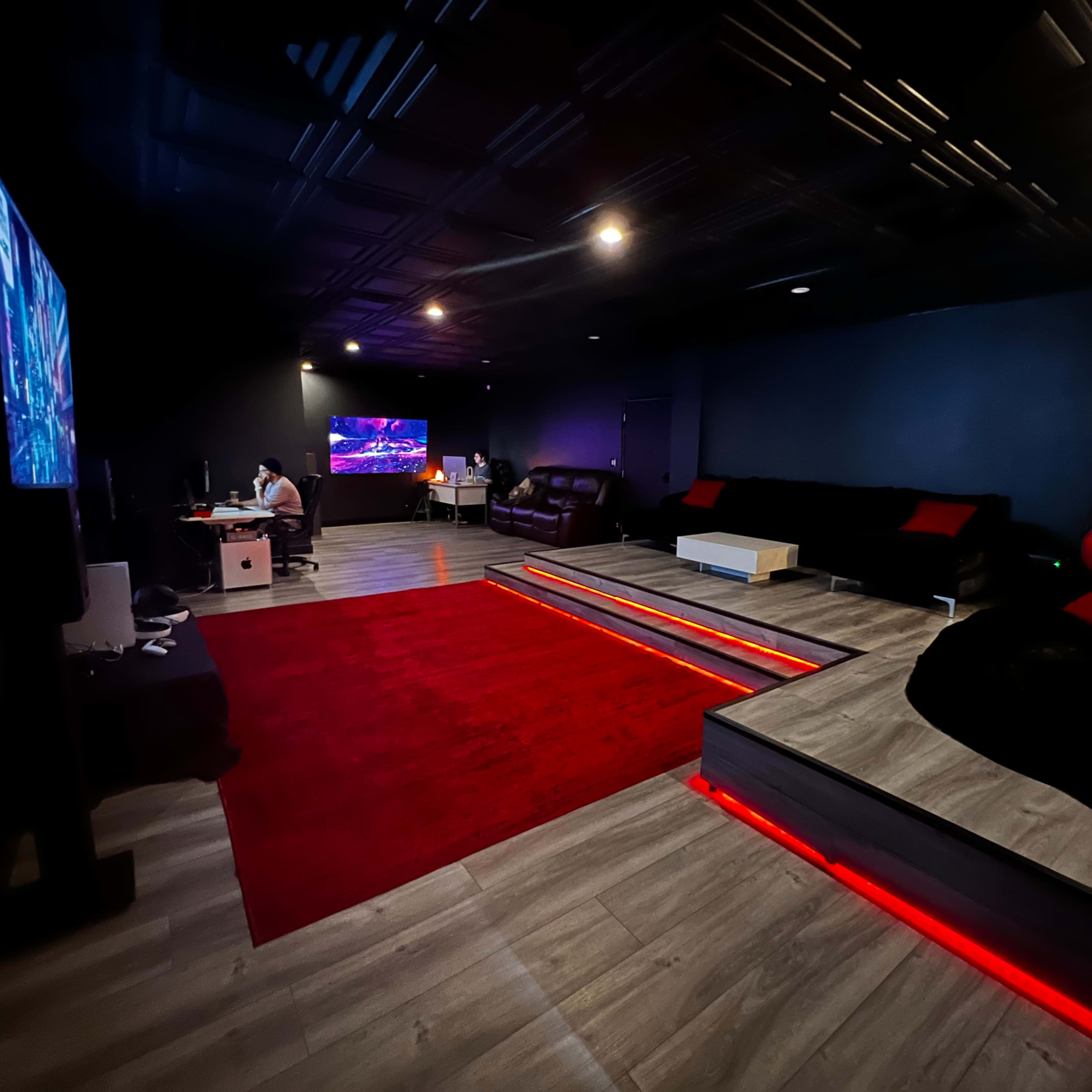 A dimly lit entertainment room featuring two large screens, a red area rug, tiered seating, and cozy seating arrangements.