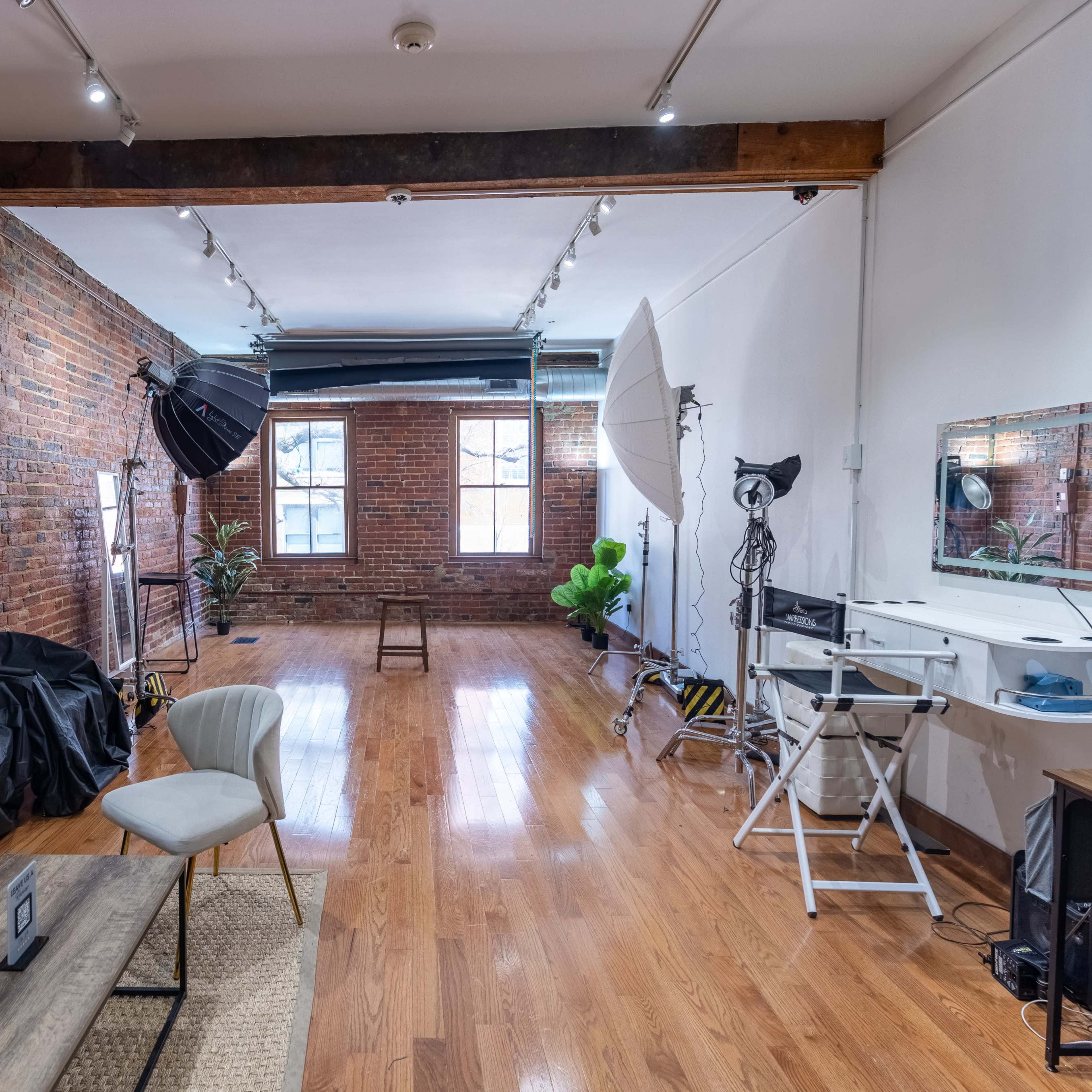 A spacious room with exposed brick walls, hardwood floors, and various photography equipment set up, including softboxes and a makeup area.