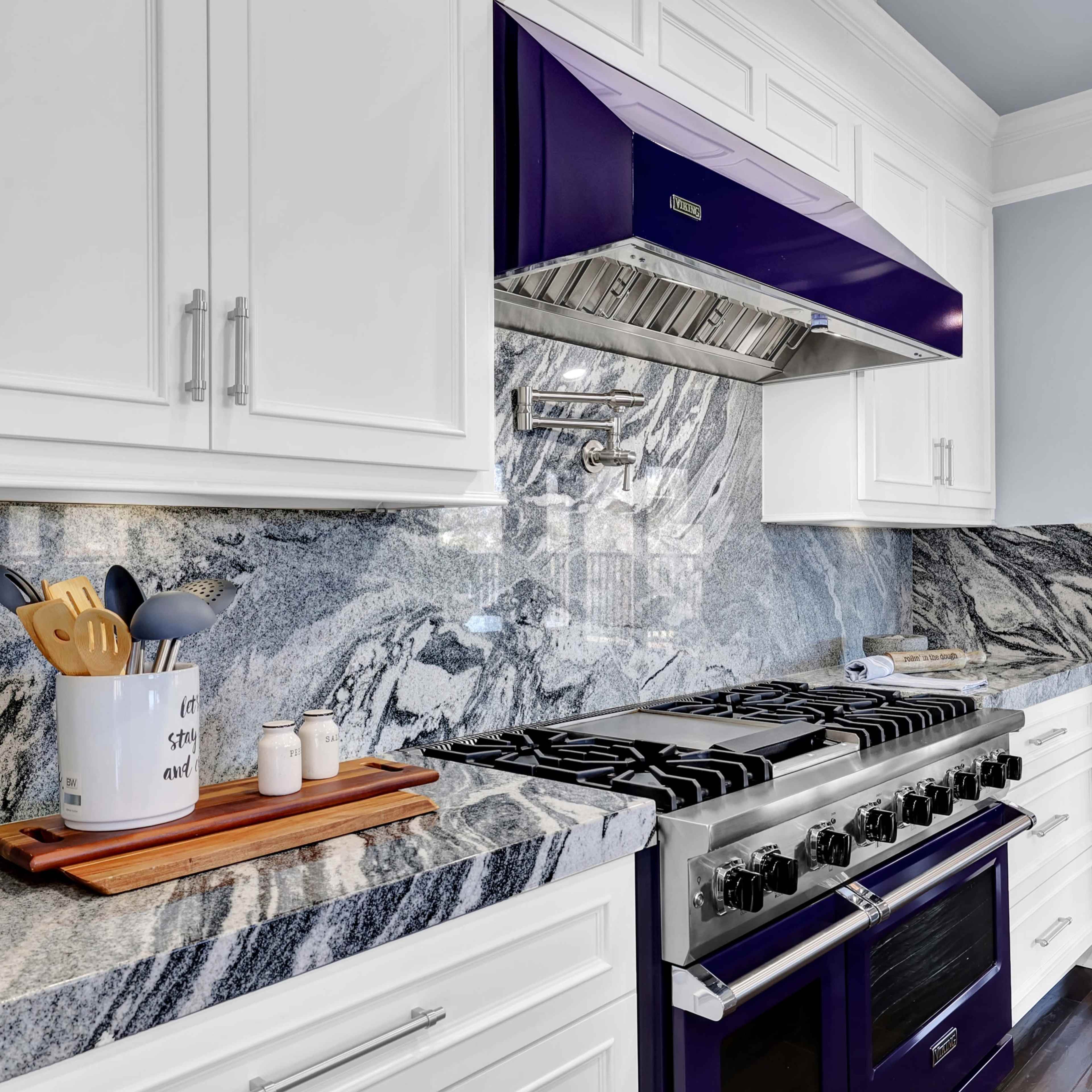 A modern kitchen featuring a stainless steel gas stove with multiple burners, a purple range hood, and white cabinetry, all set against a marble countertop.