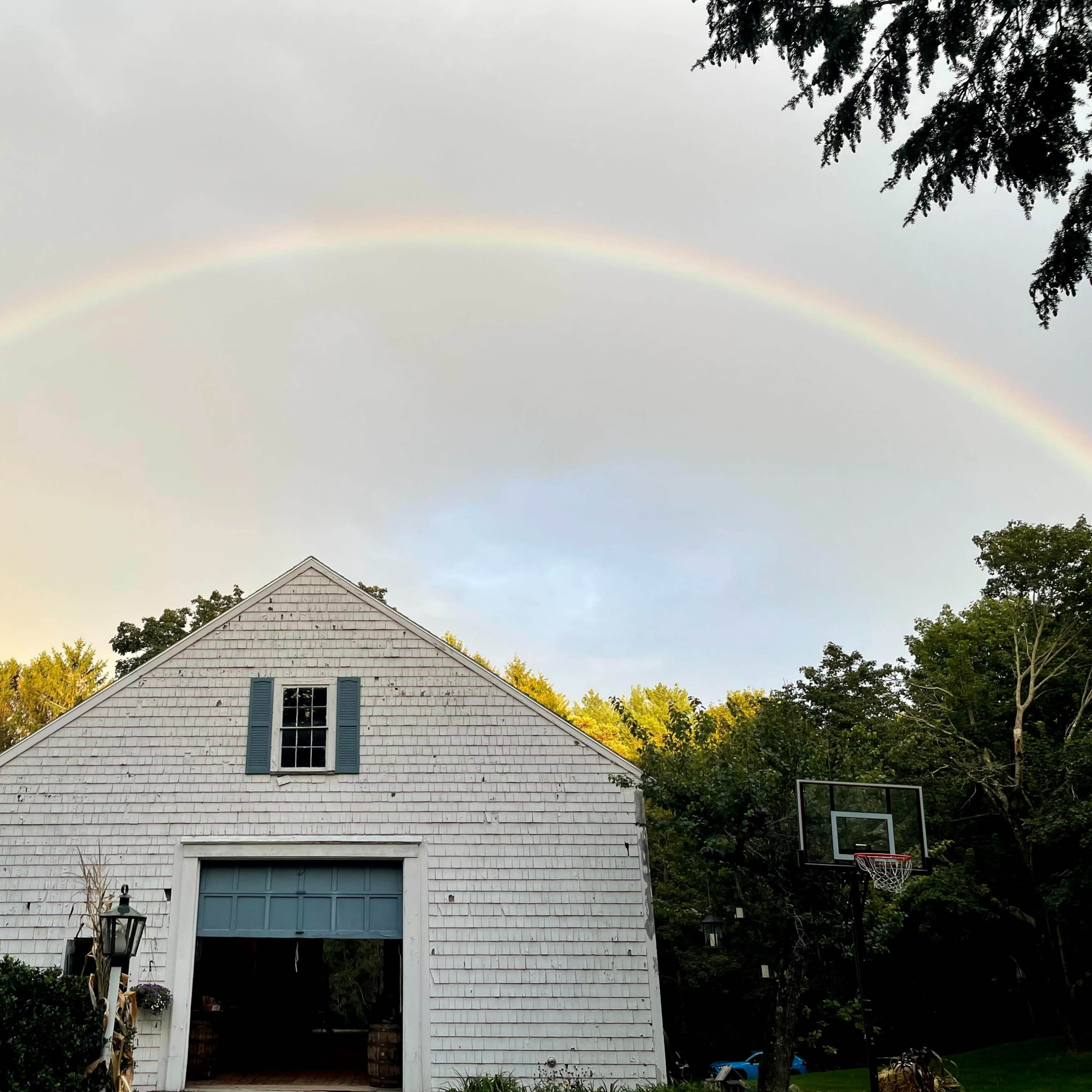 A rainbow arcs over a white barn-style garage surrounded by trees and a basketball hoop.
