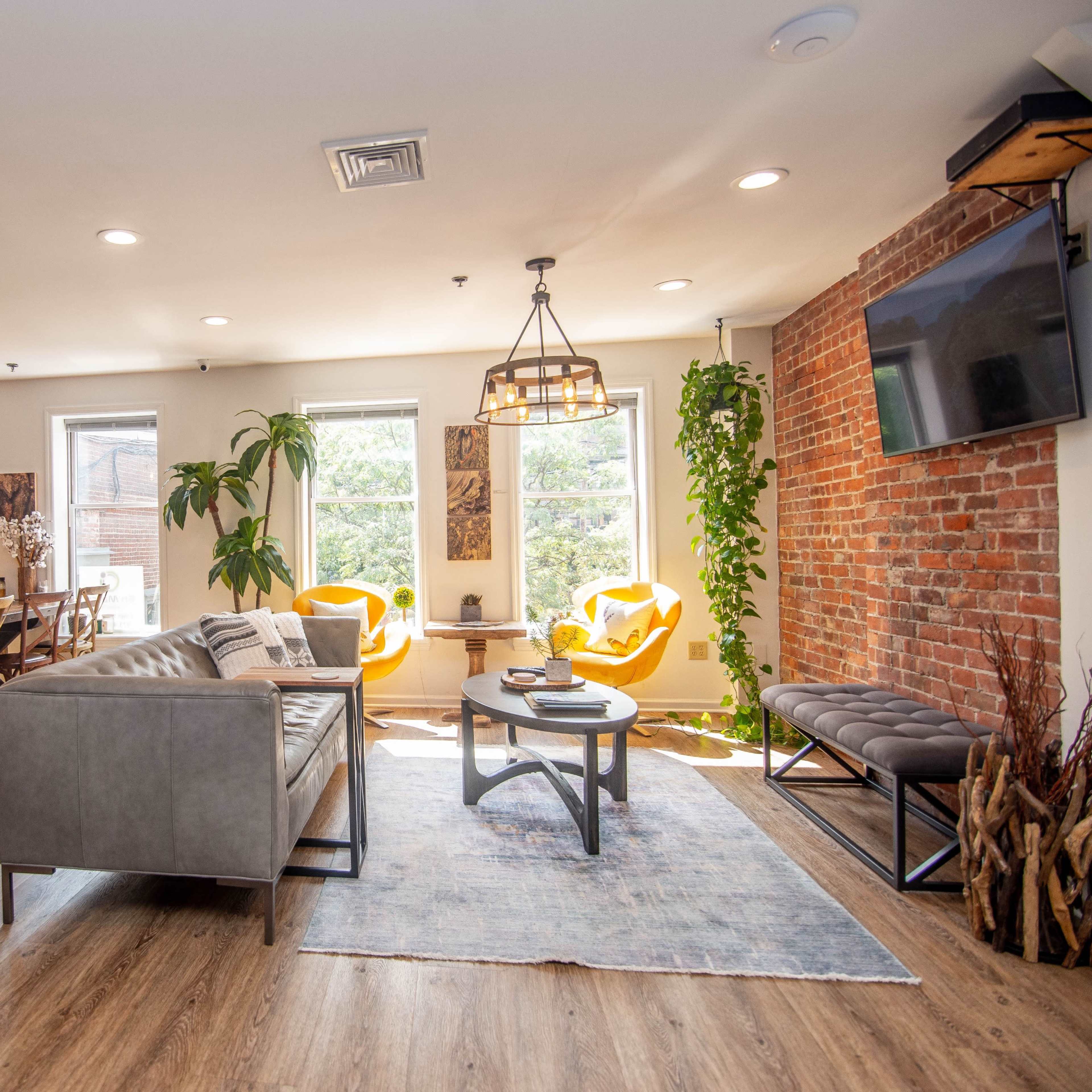 A modern lounge area with a gray sofa, a wooden dining table, bright yellow armchairs, and brick walls, illuminated by natural light from the windows.