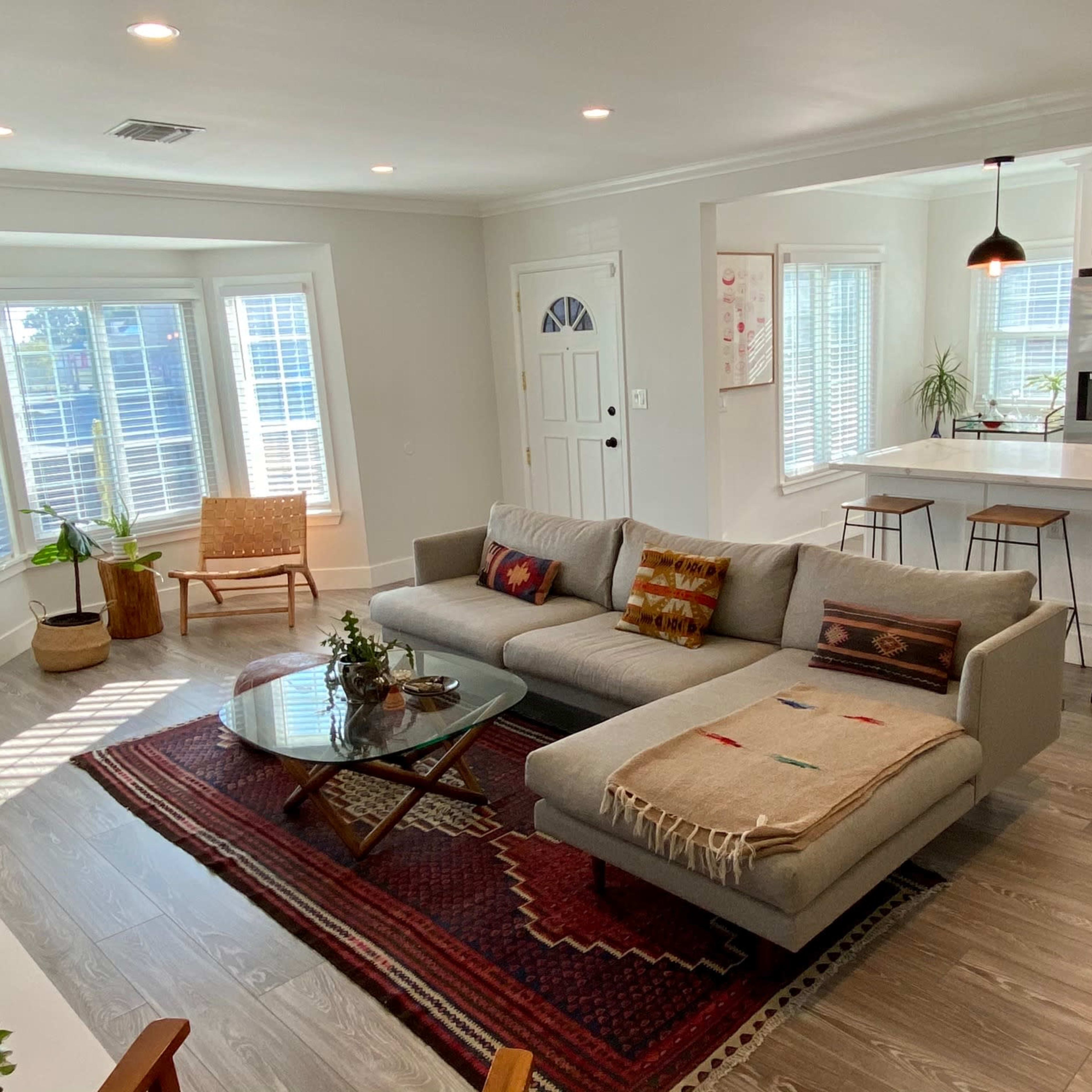 A modern living room with a gray sectional sofa, a round glass coffee table, and a dining area featuring bar stools and a kitchen in the background.