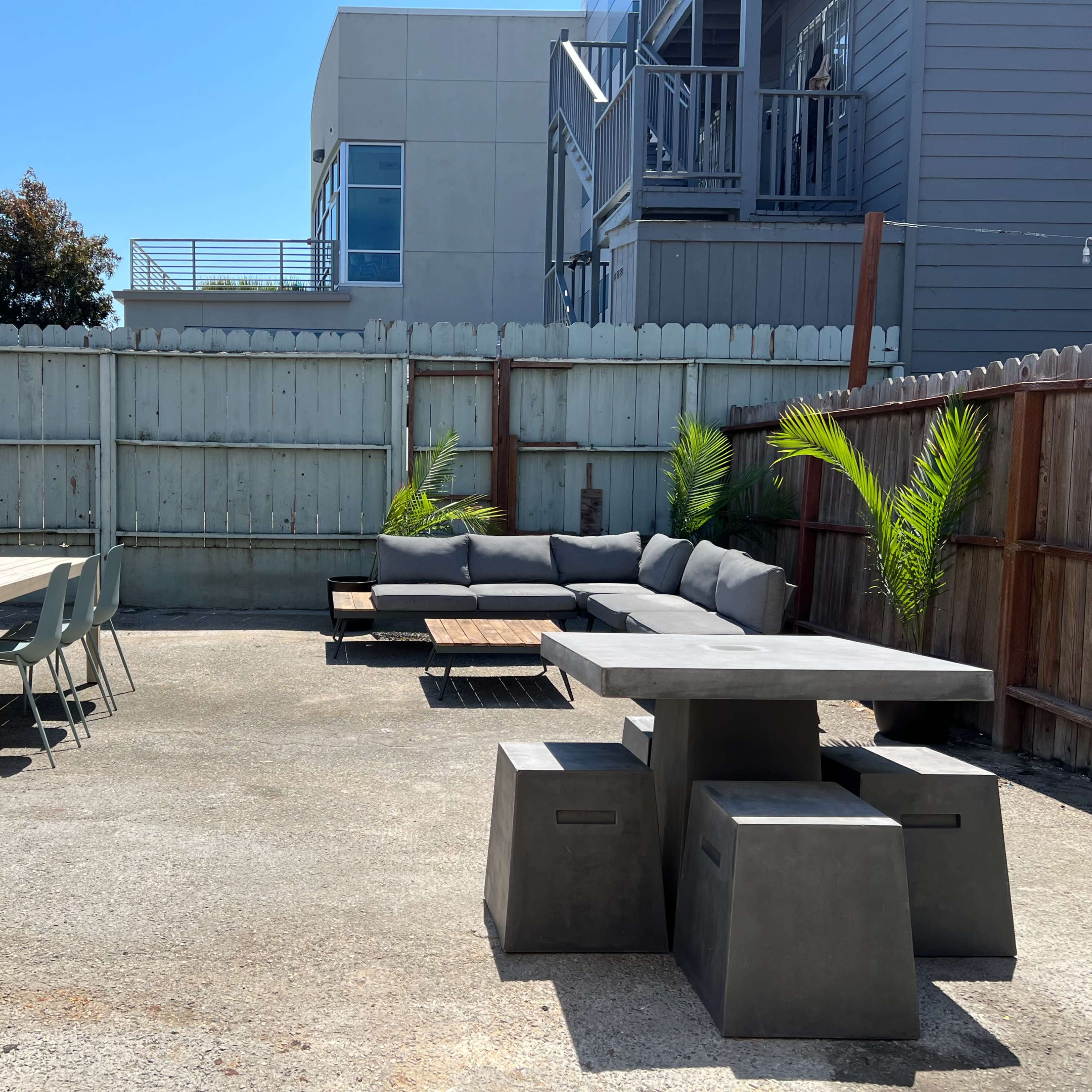 A patio area features a concrete table with seating, a gray sectional couch, and a dining table surrounded by chairs, all enclosed by a wooden fence.