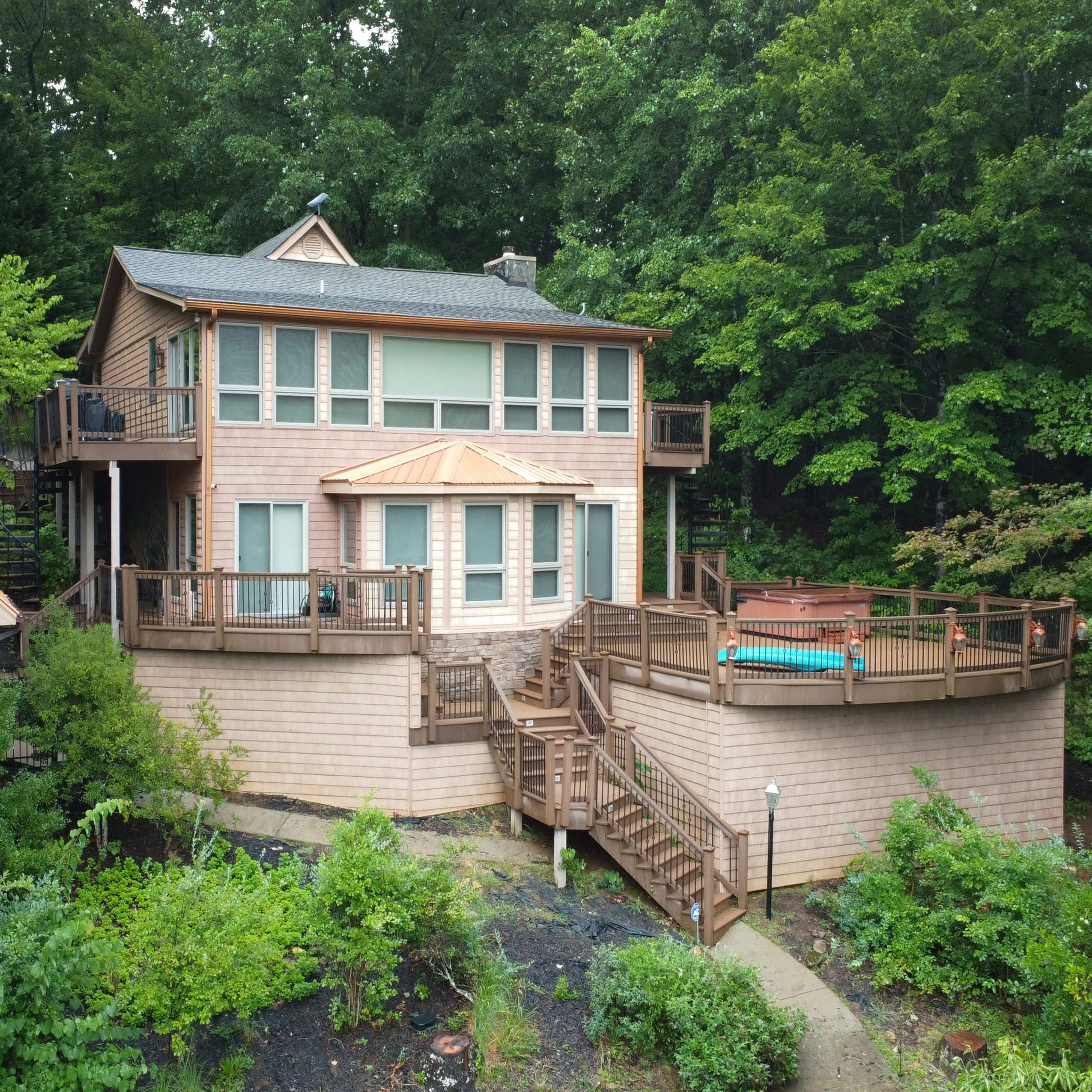 A two-story wooden house with a large deck, surrounded by greenery and trees.