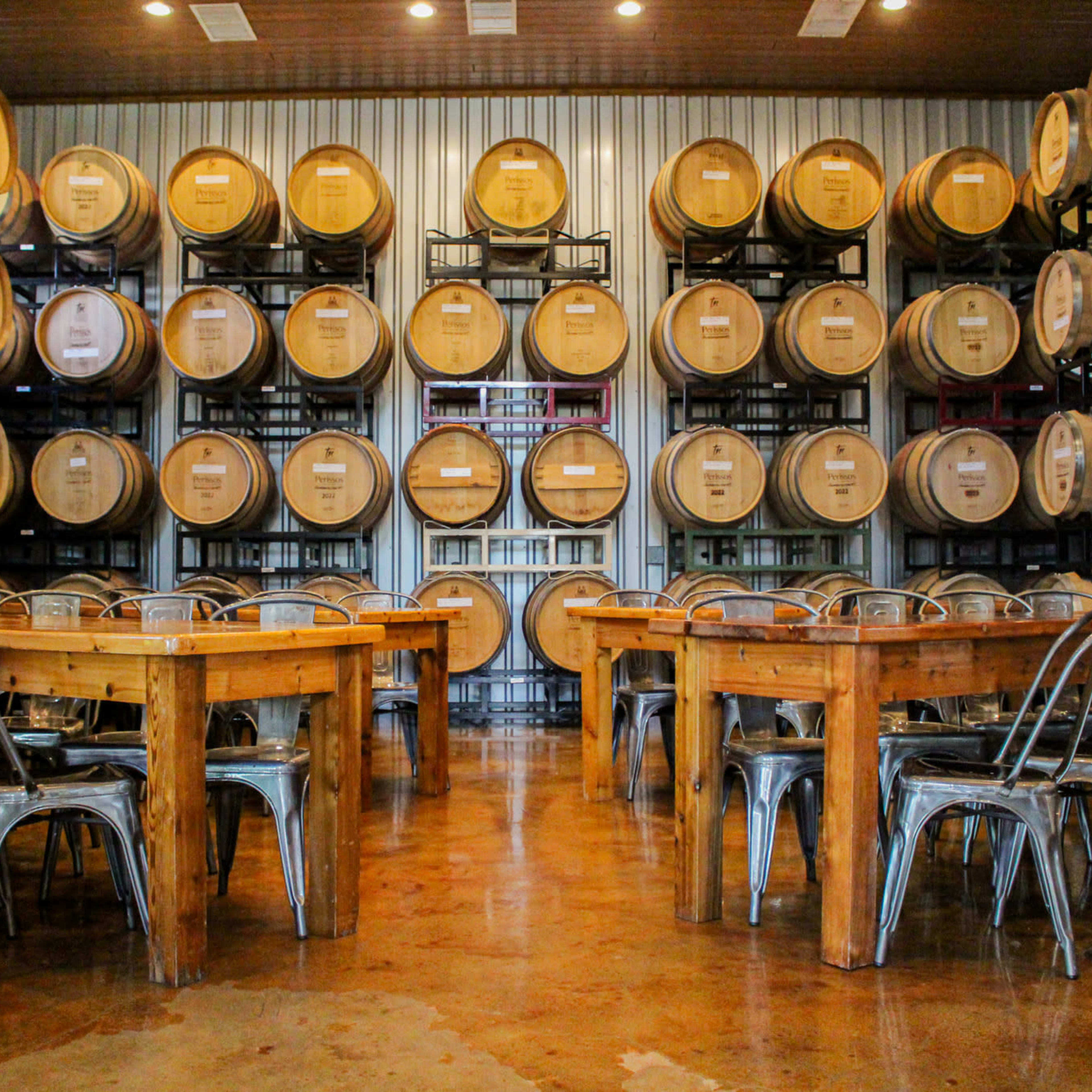 A spacious tasting room features wooden tables surrounded by metal chairs, with rows of wine barrels stacked against the wall.