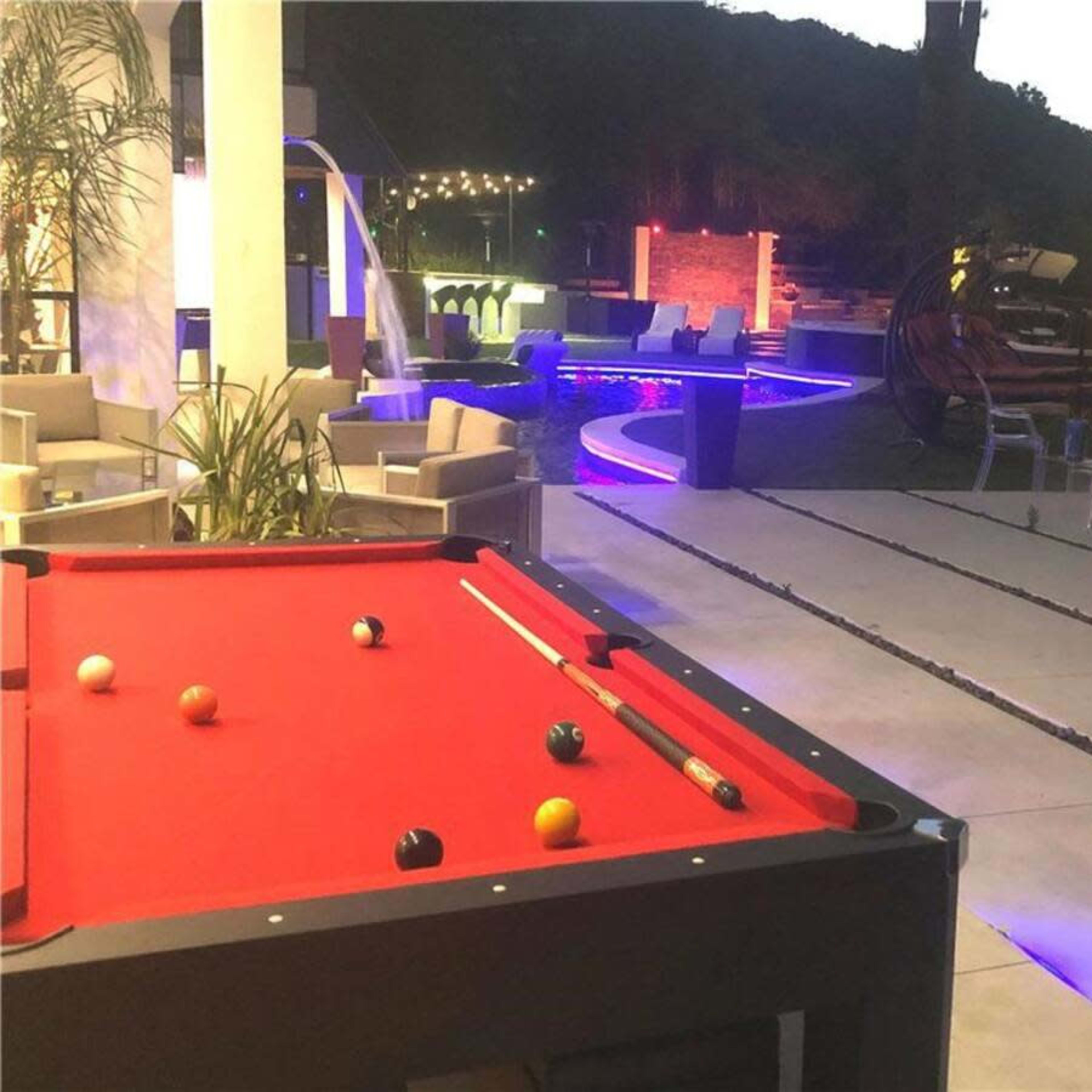 The scene shows a pool table with colorful balls in the foreground, and a well-lit outdoor area with a swimming pool and lounge seating in the background.