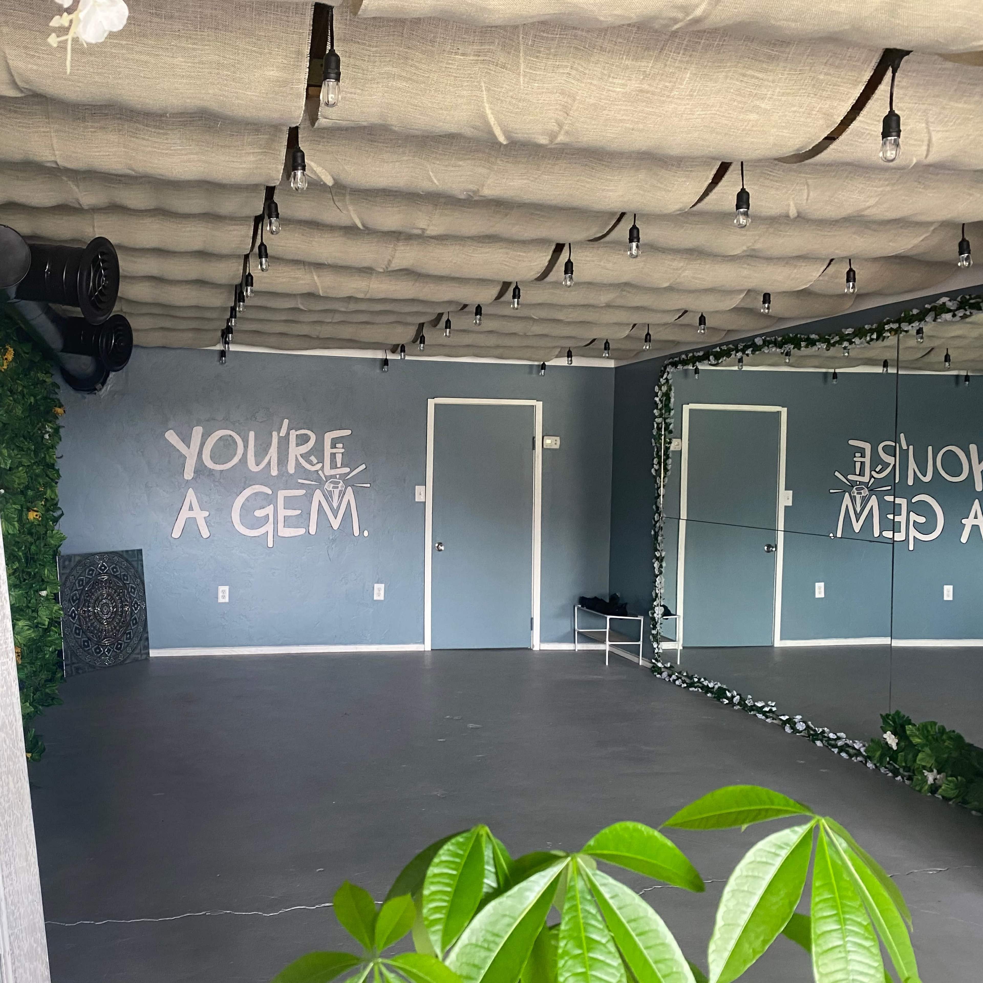 An empty room with a mirrored wall and a message on the wall that reads "YOU'RE A GEM," surrounded by greenery and soft lighting.