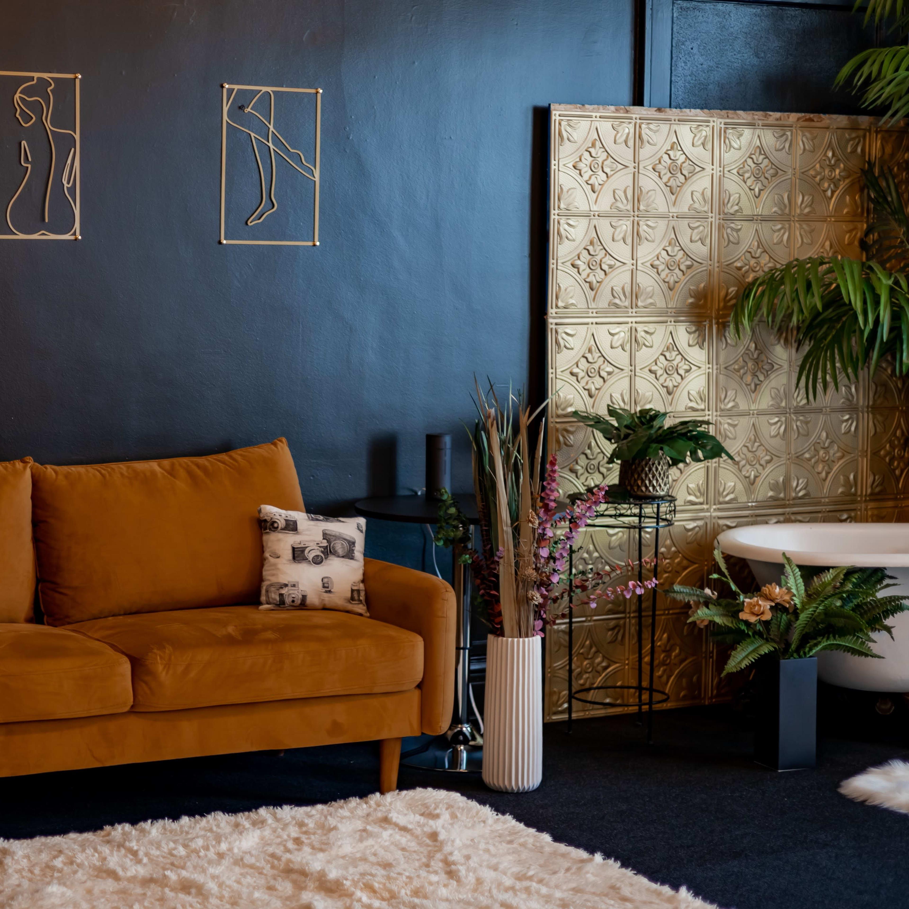 A cozy living area featuring a brown suede sofa, gold wall art, a small white rug, and a bathtub surrounded by plants.