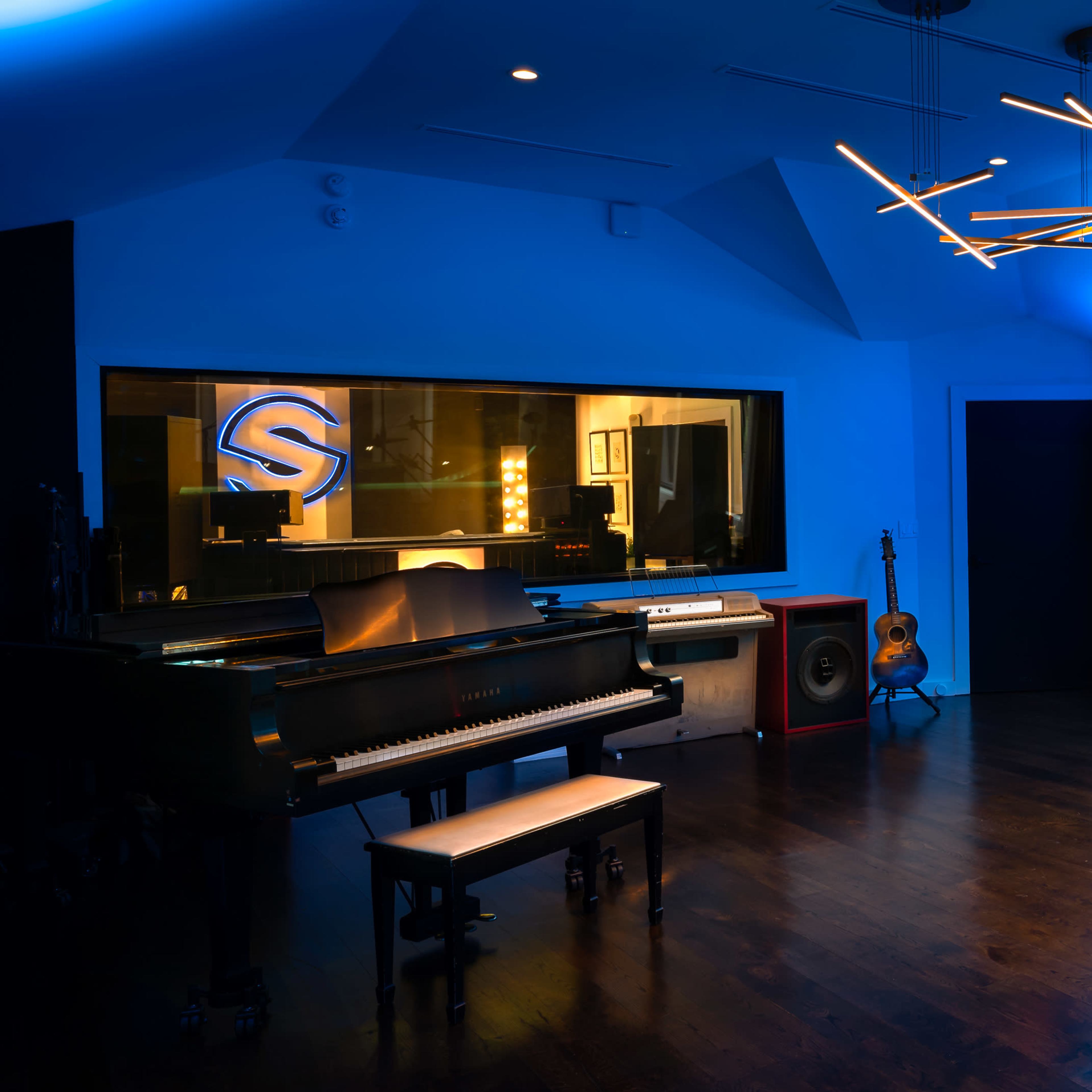 A modern music studio features a grand piano, a drum set, and a guitar, illuminated by blue lighting and a geometric chandelier.