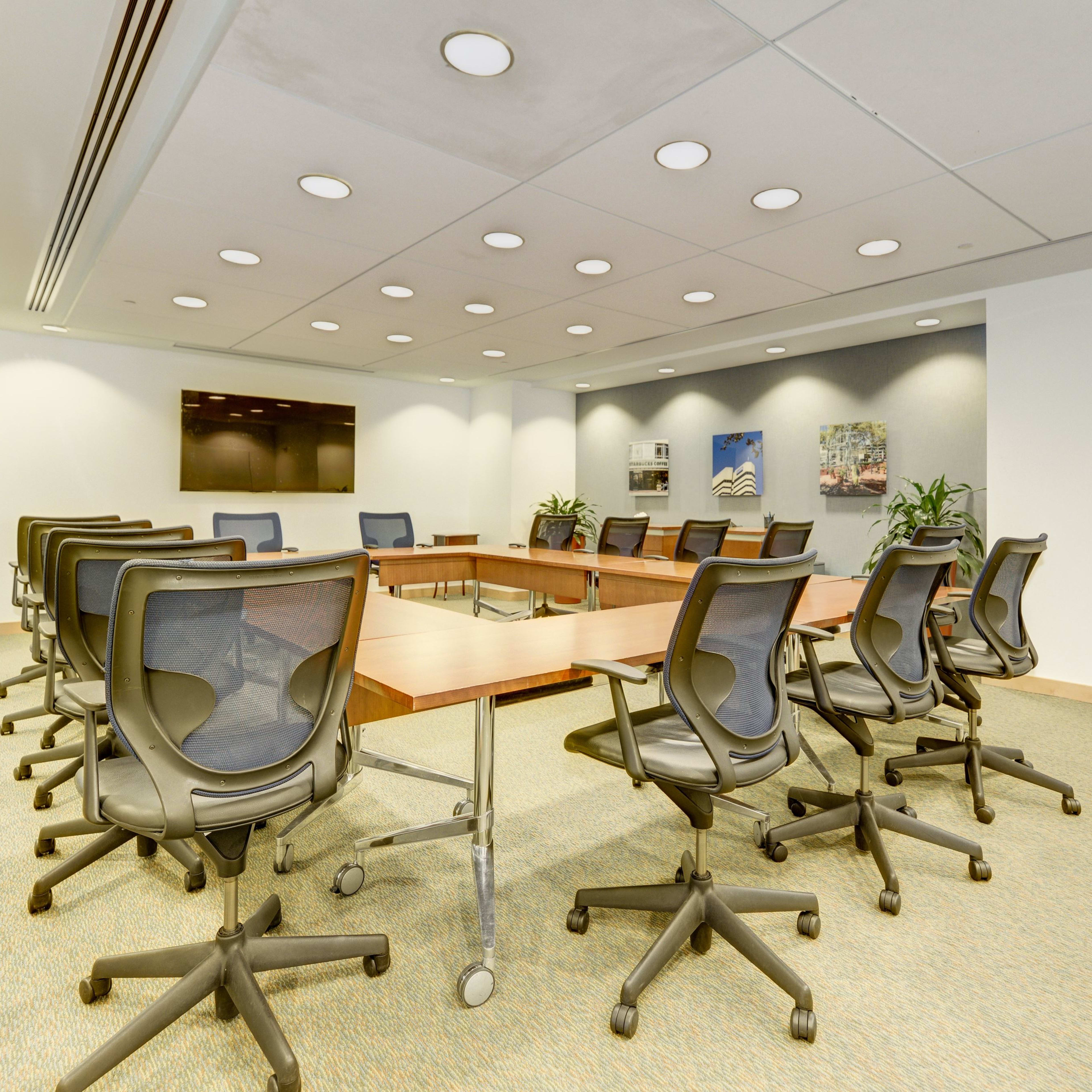 A spacious conference room features a large wooden table surrounded by black swivel chairs, with potted plants and a wall-mounted screen at one end.