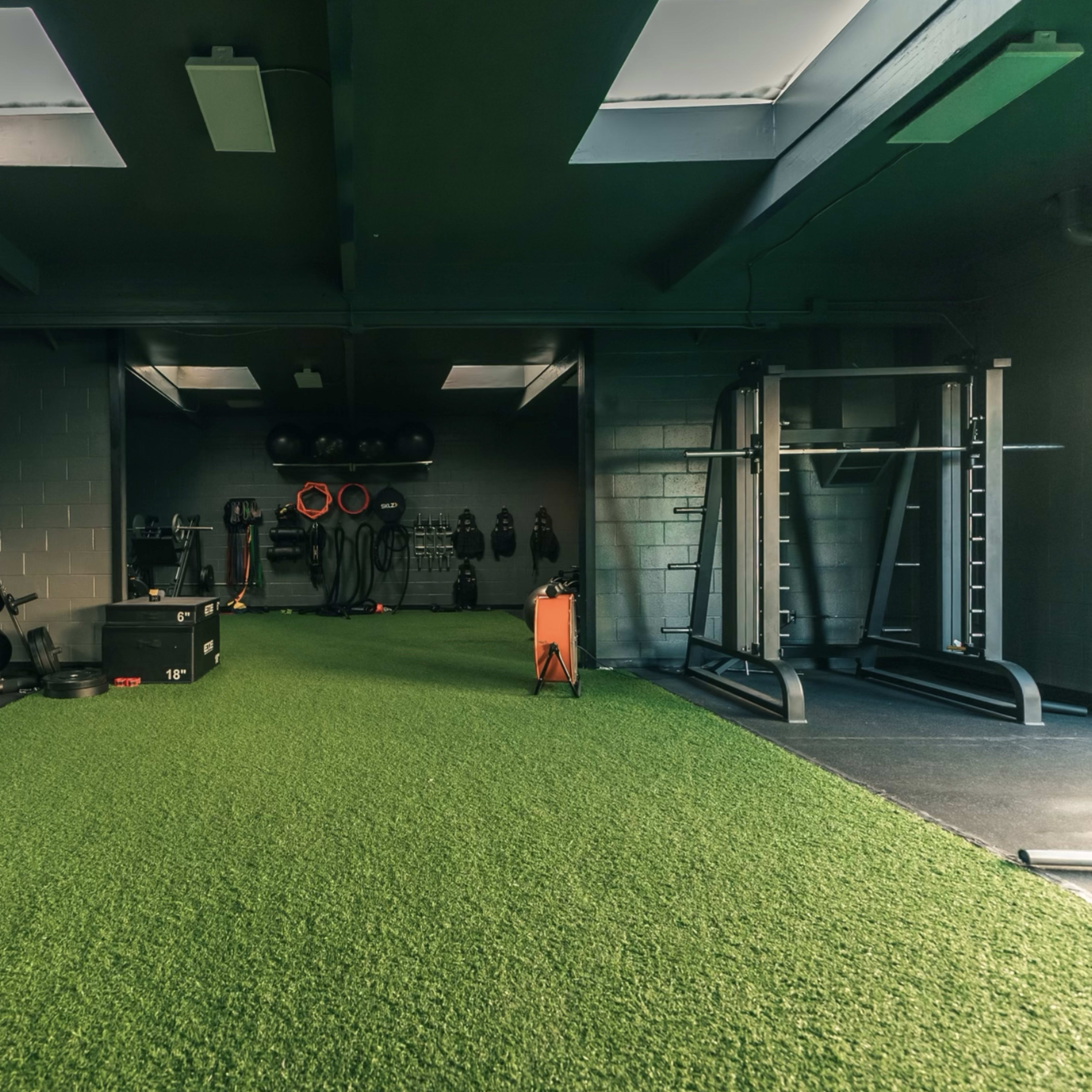 A fitness gym interior features artificial grass flooring, various exercise equipment, and weights arranged along the walls.