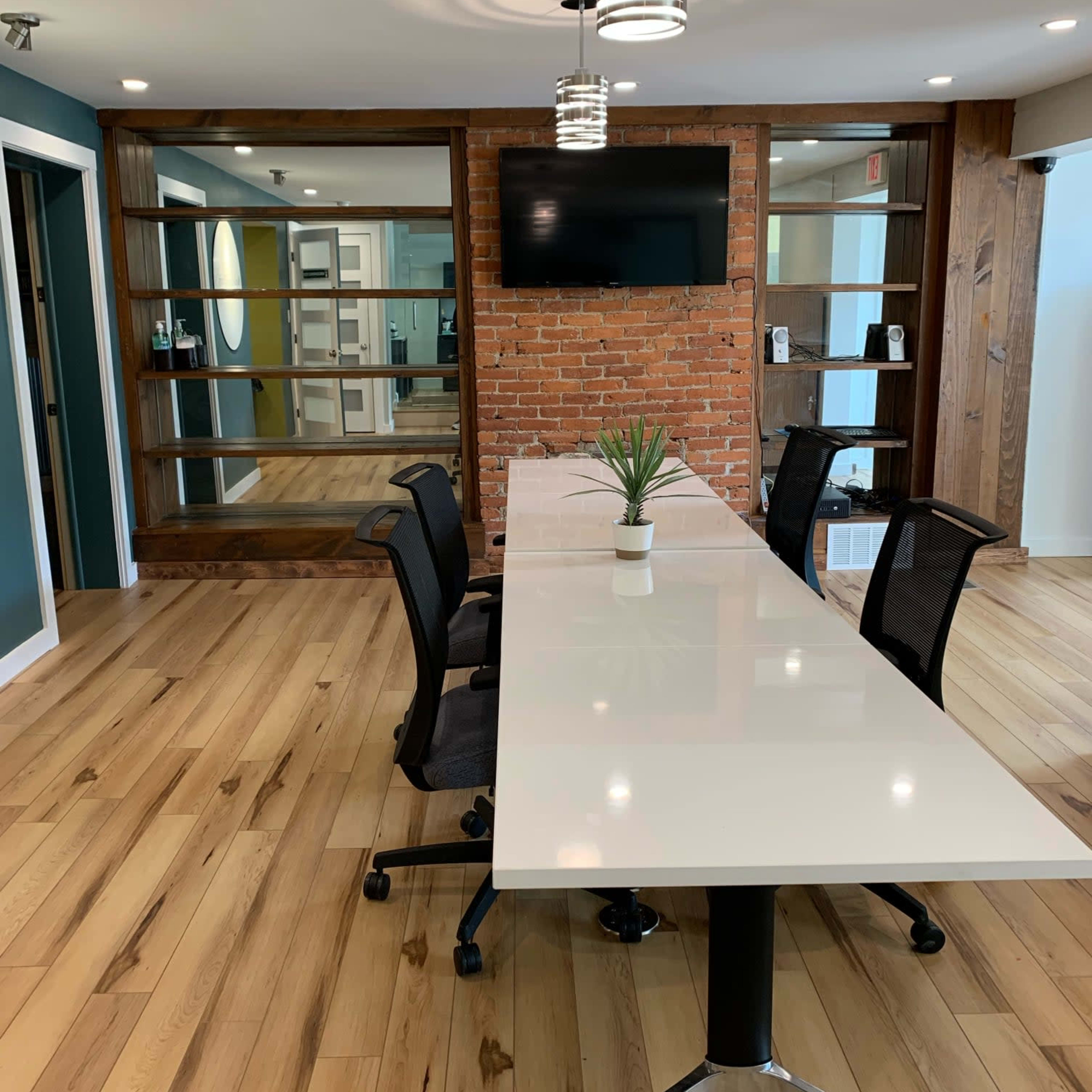 A modern conference room with a long white table, four black chairs, a television mounted on a brick wall, and glass shelving units.