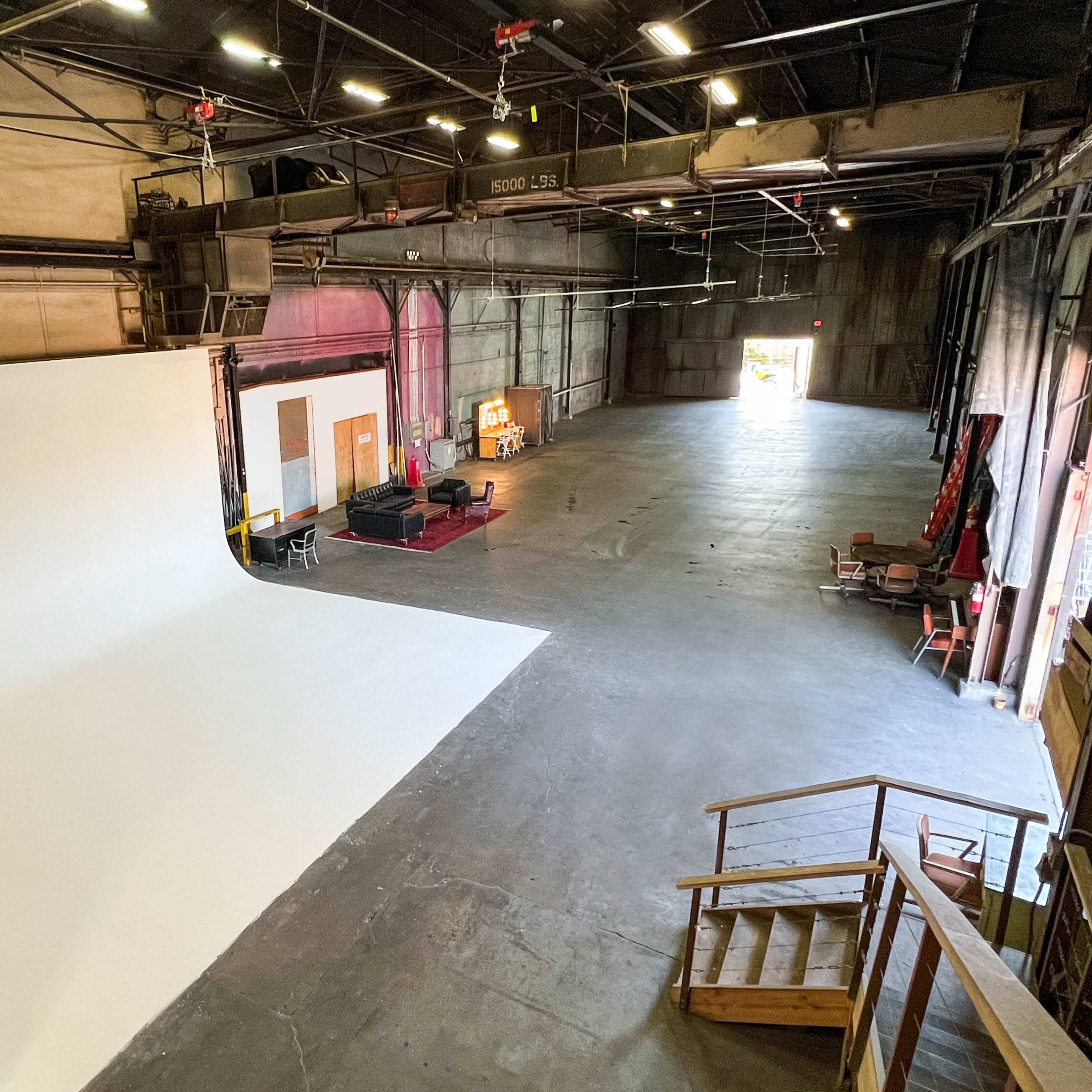 A spacious studio with a large white backdrop, industrial lighting, and various pieces of furniture in a well-lit area.