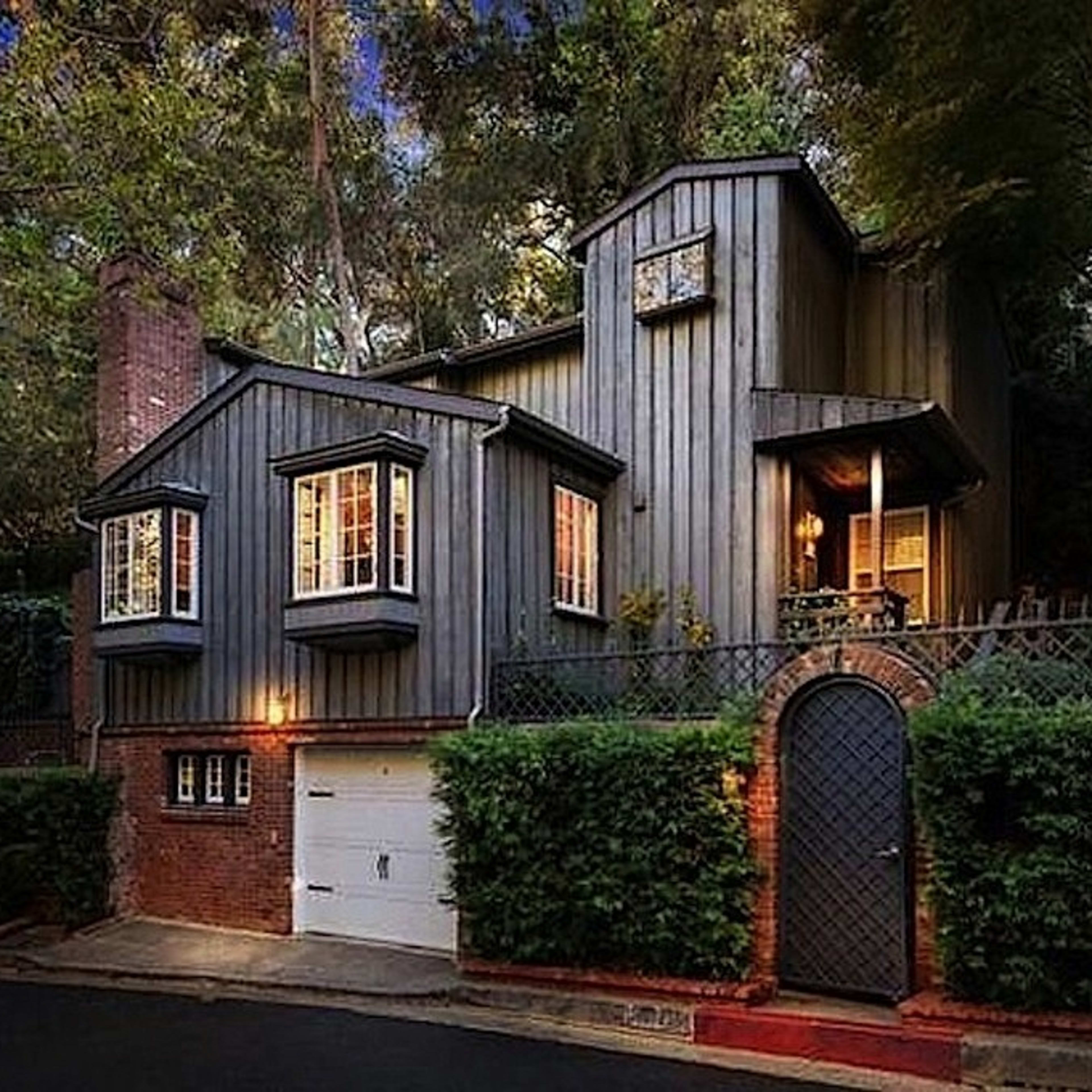 A two-story wooden house with large windows and a brick garage is surrounded by trees and greenery.