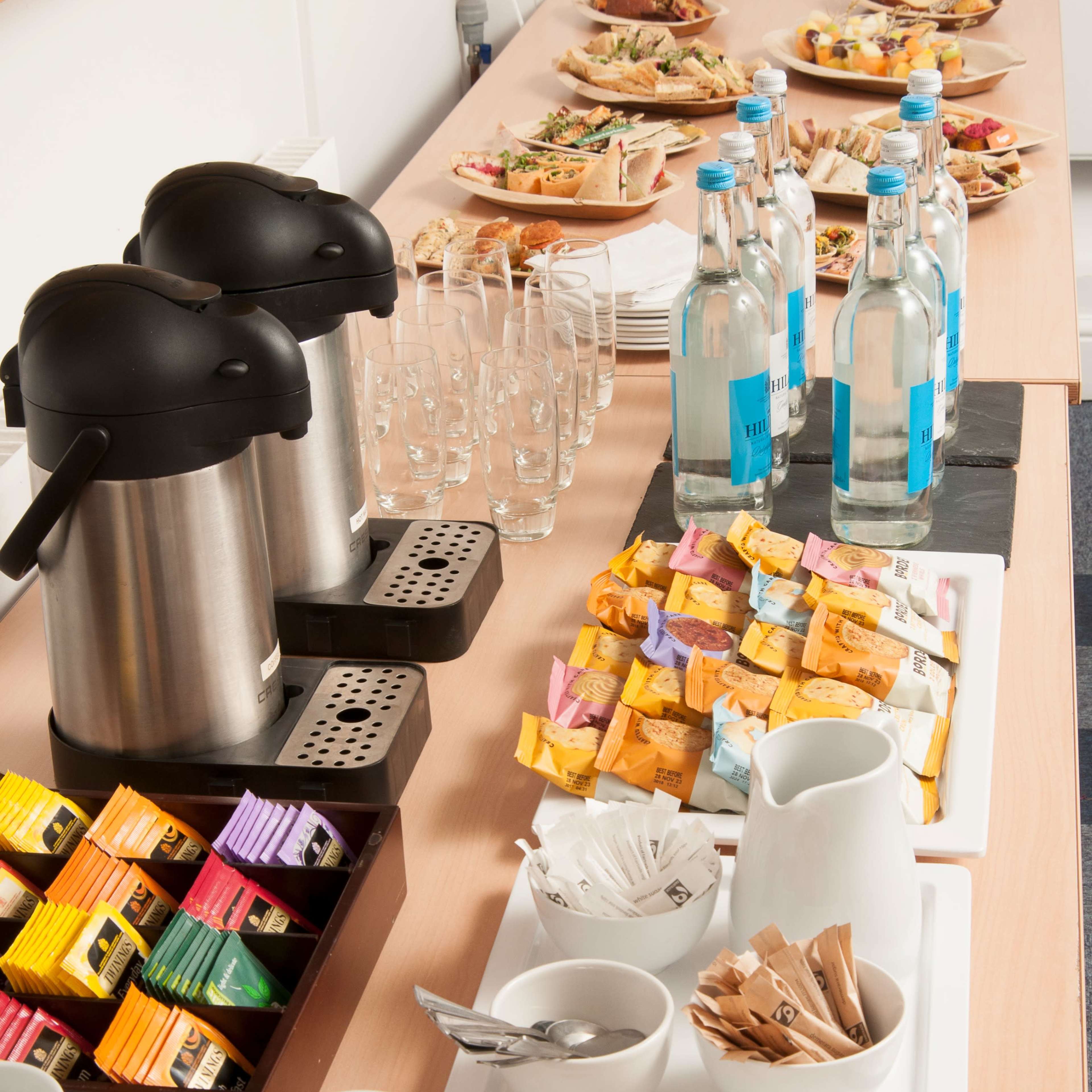 A refreshment table with coffee thermoses, bottles of water, assorted tea bags, cups, and a variety of snacks on platters.