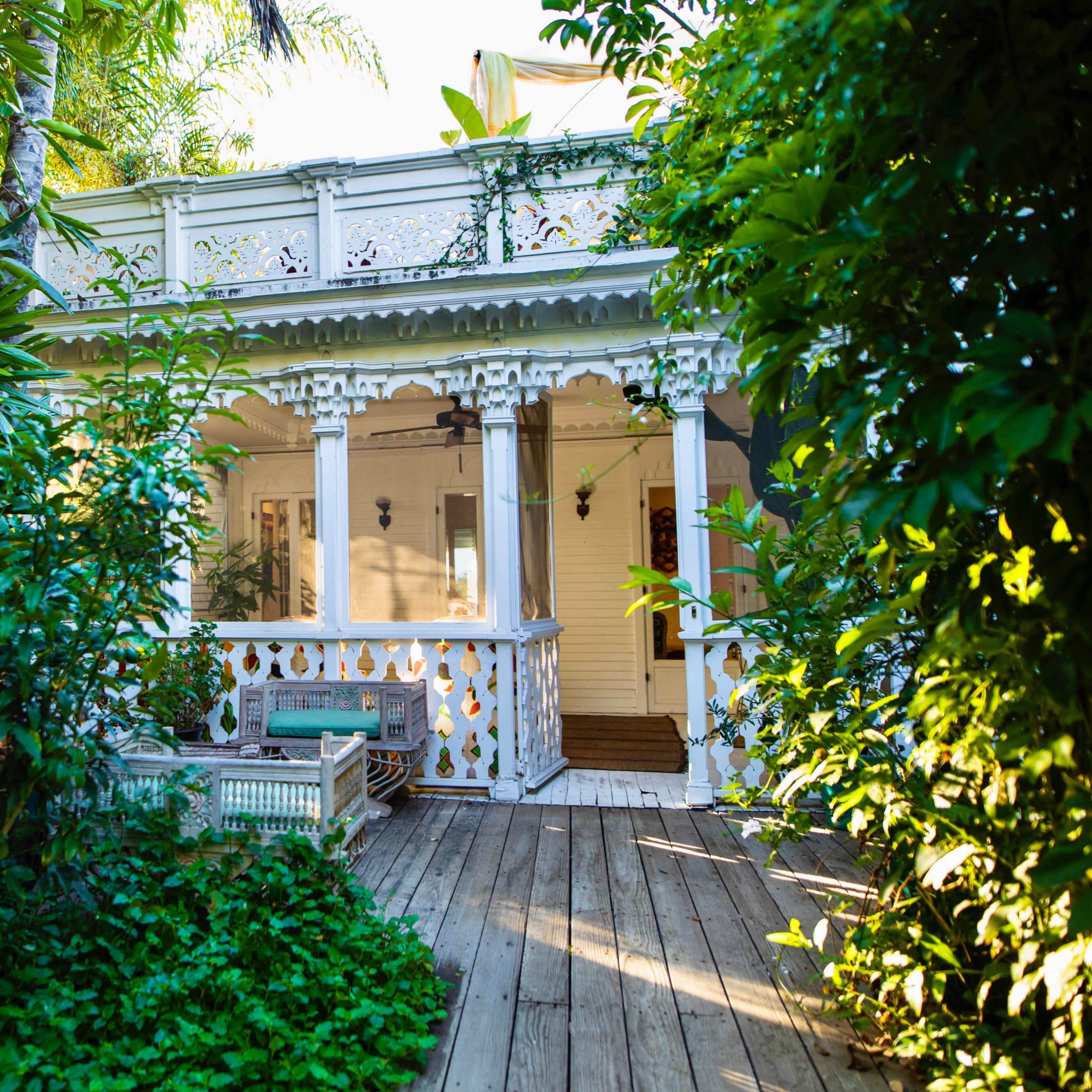 A white, Victorian-style house is nestled among lush greenery, with a wooden deck leading to its entrance.