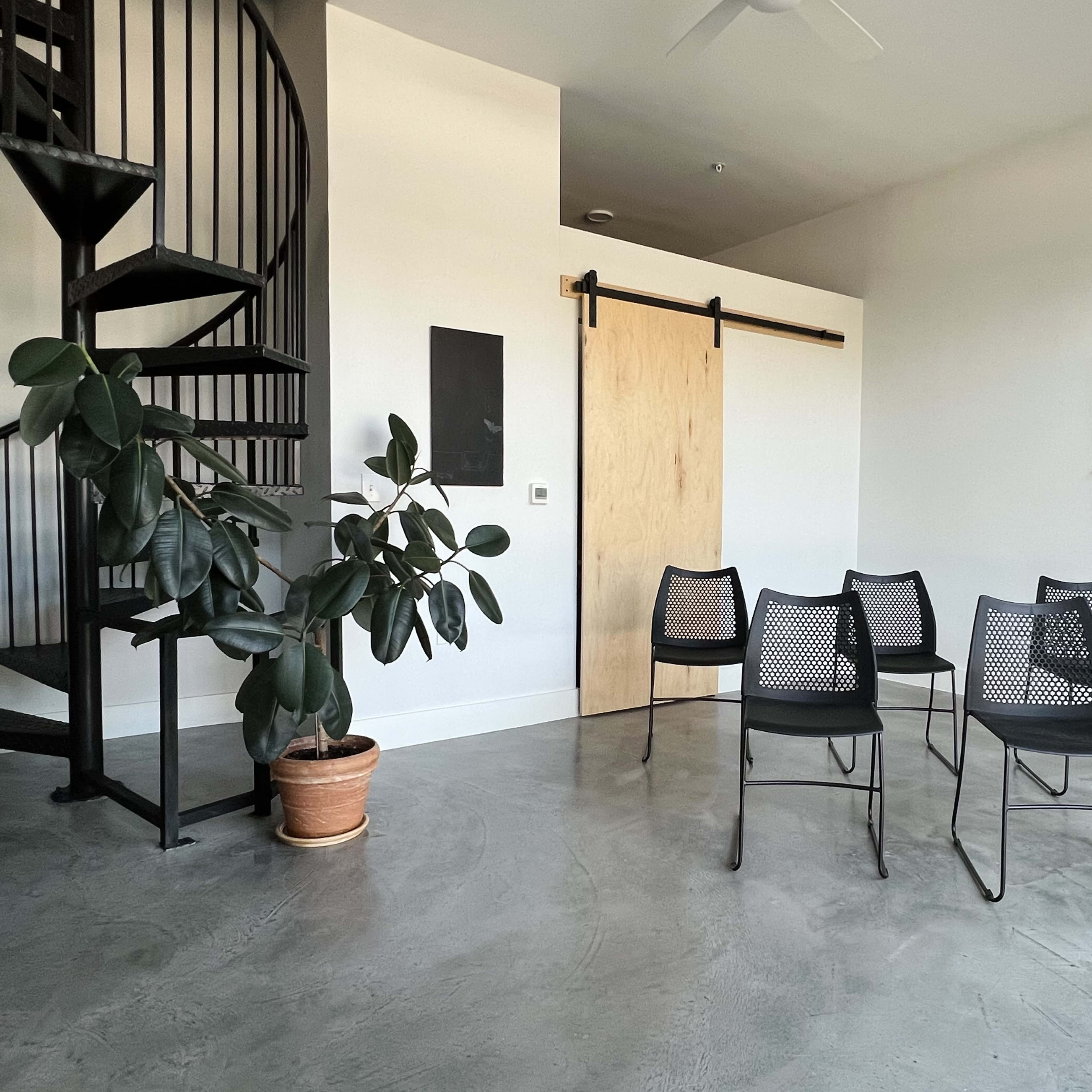 A modern, minimalist room with a spiral staircase, a potted plant, and six black chairs arranged around a central area.