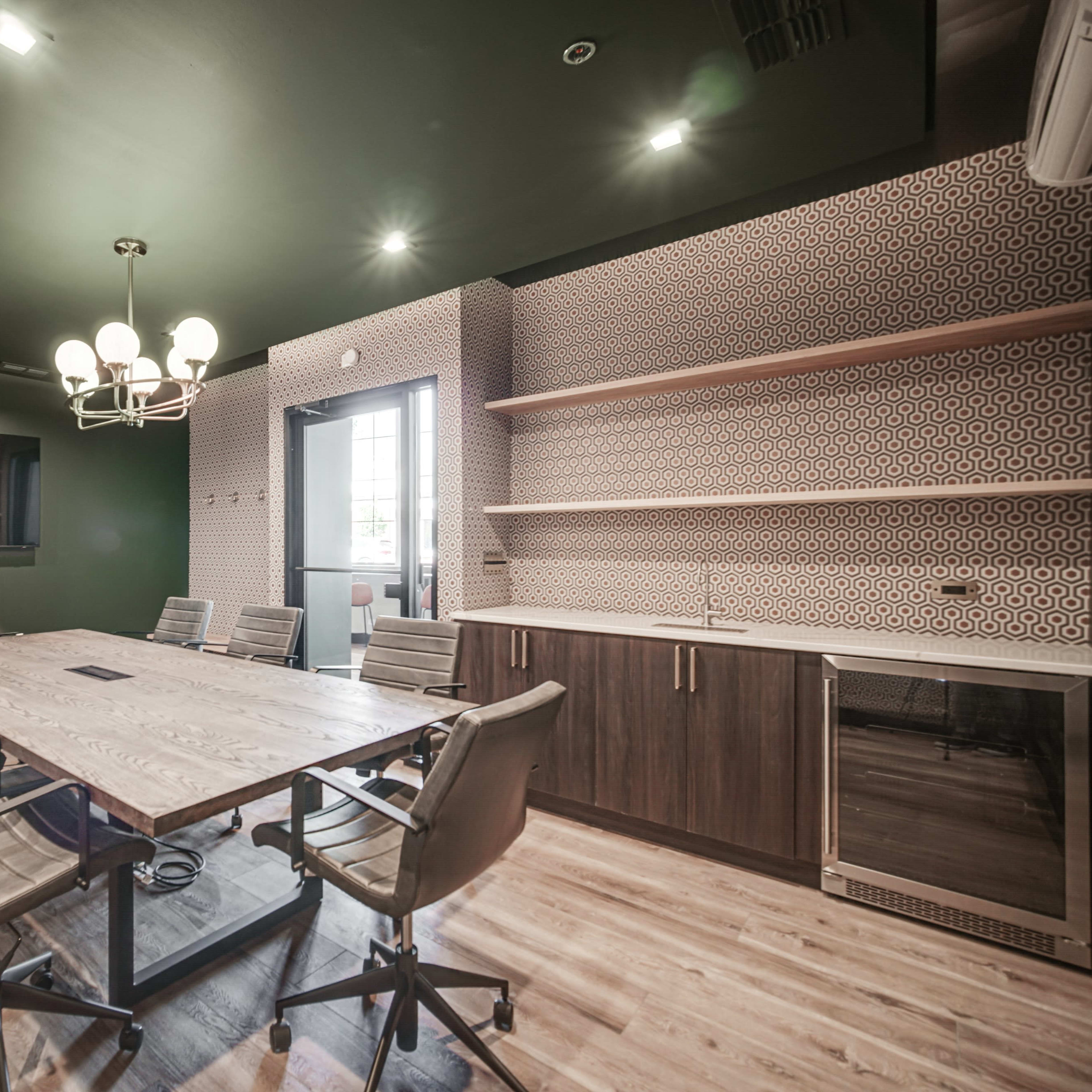 A modern conference room features a large wooden table surrounded by ergonomic chairs, with a wall-mounted screen and a kitchenette in the background.