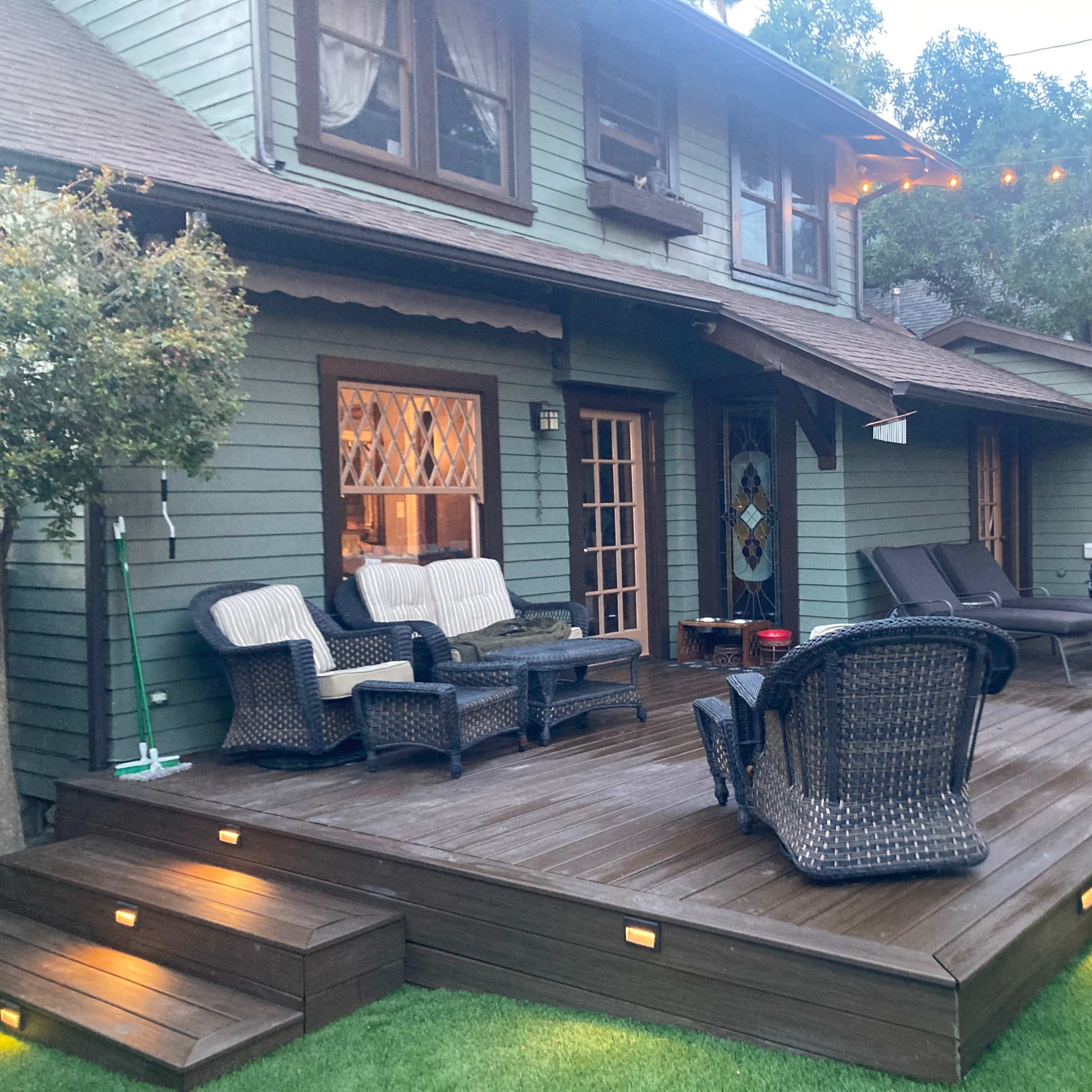 A two-story house with a wooden deck featuring lounge chairs, outdoor seating, and a hot tub, surrounded by greenery and lighted string decorations.