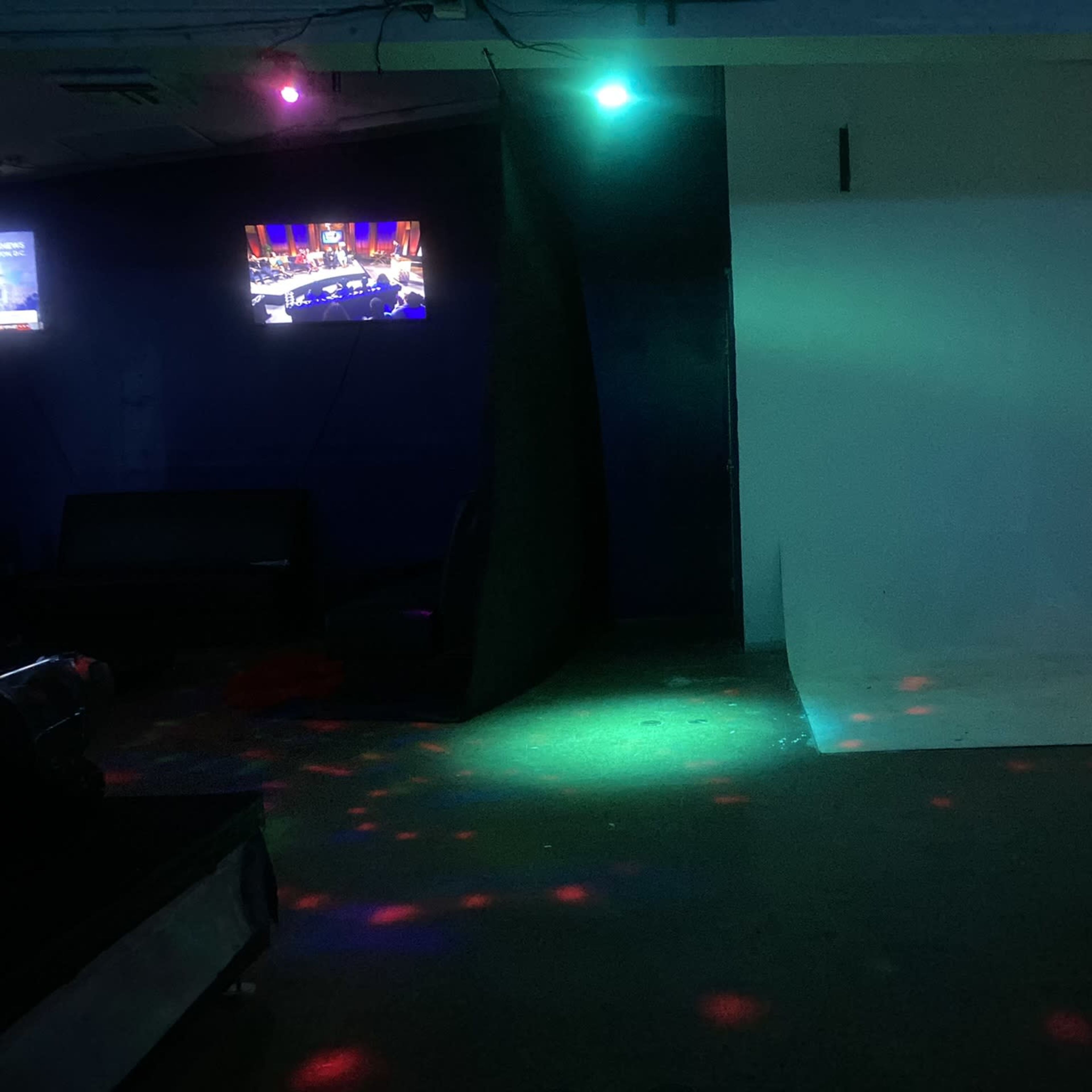 A dimly lit room features two televisions on one wall, a black partition, and a blank white wall illuminated by colored lights.