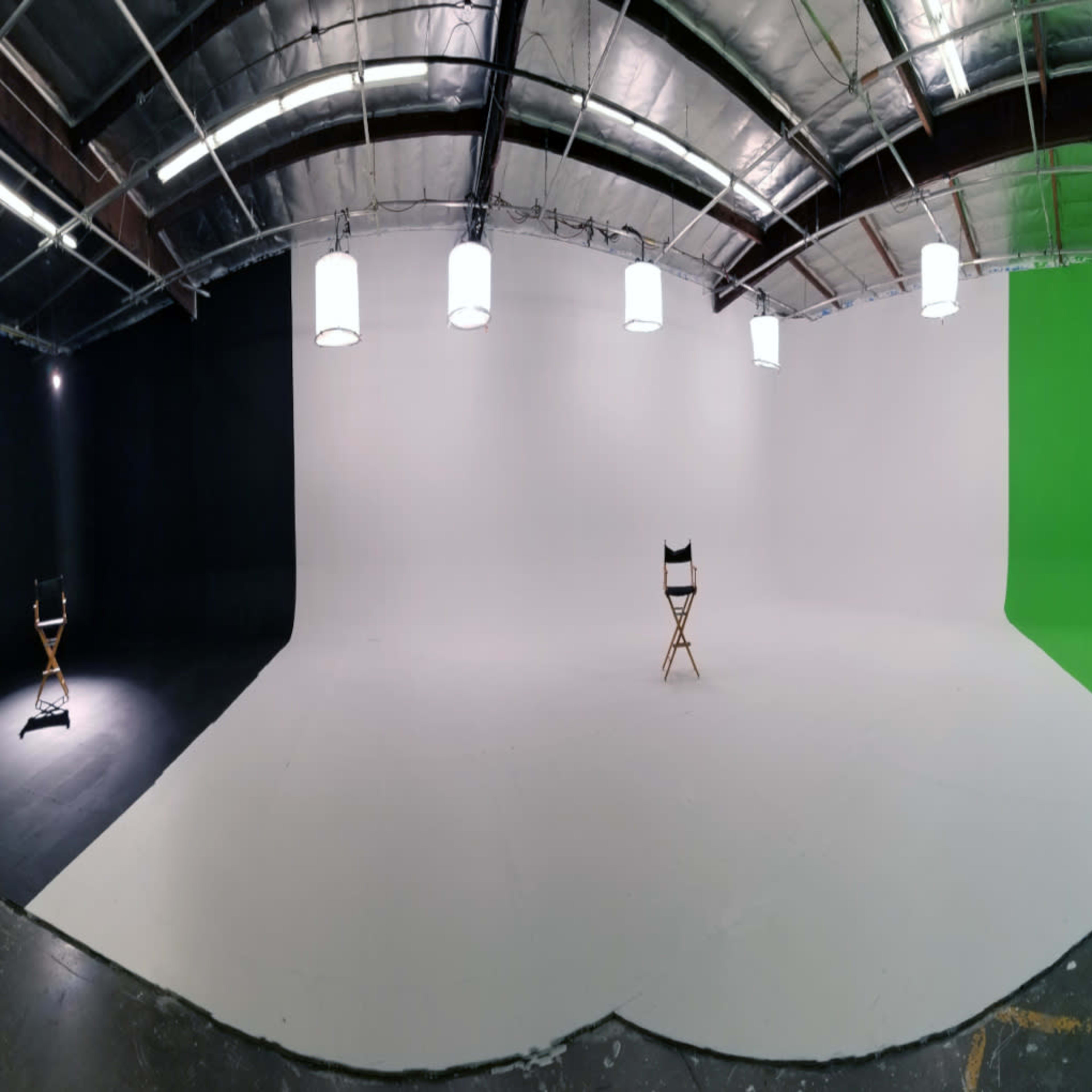 A large, empty studio space with a black wall on one side, a white wall in the center, and a green wall on the opposite side, all featuring a single wooden director's chair positioned in front of the white backdrop.