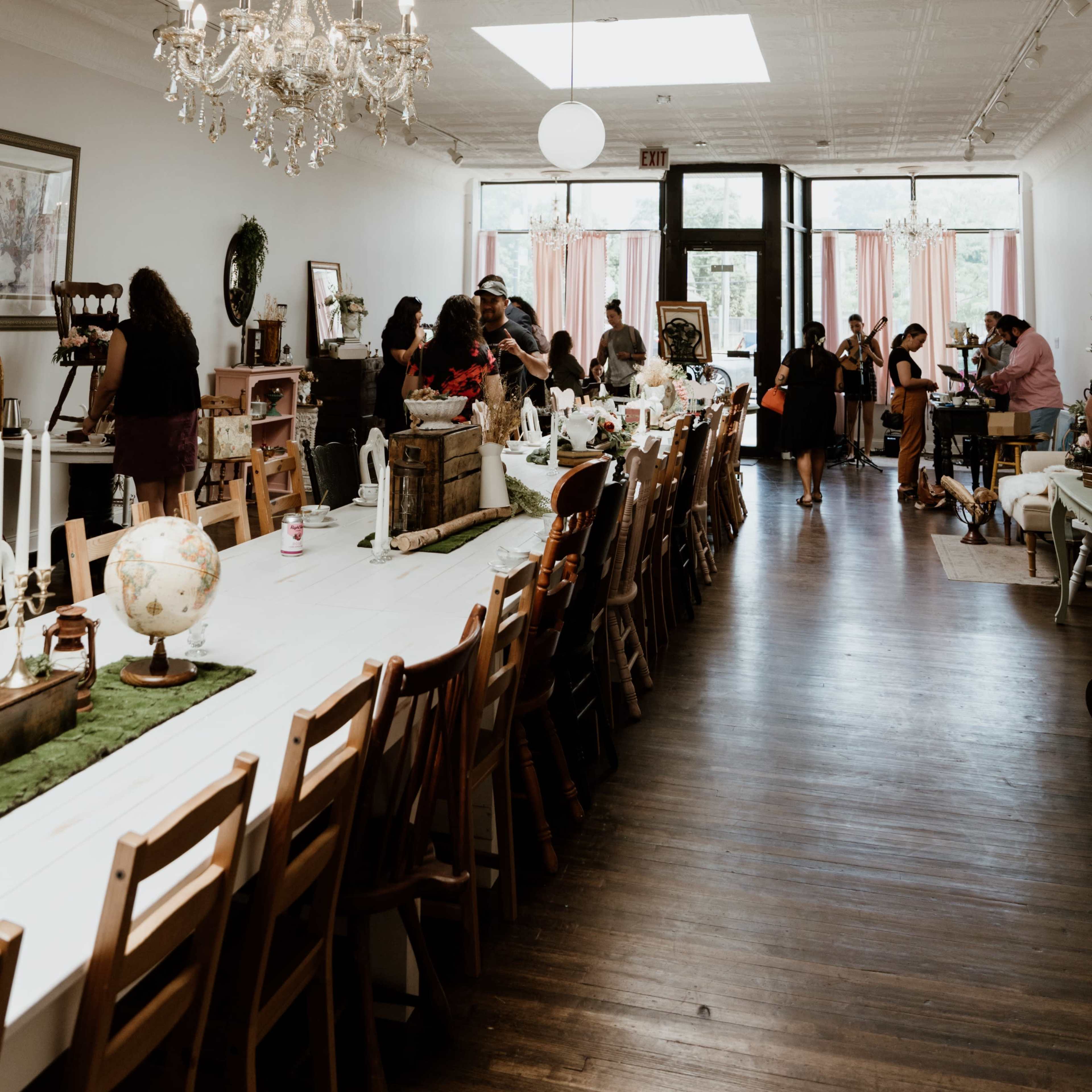 A spacious room with a long dining table and a chandelier, filled with people engaged in activities, while sunlight streams through large windows.