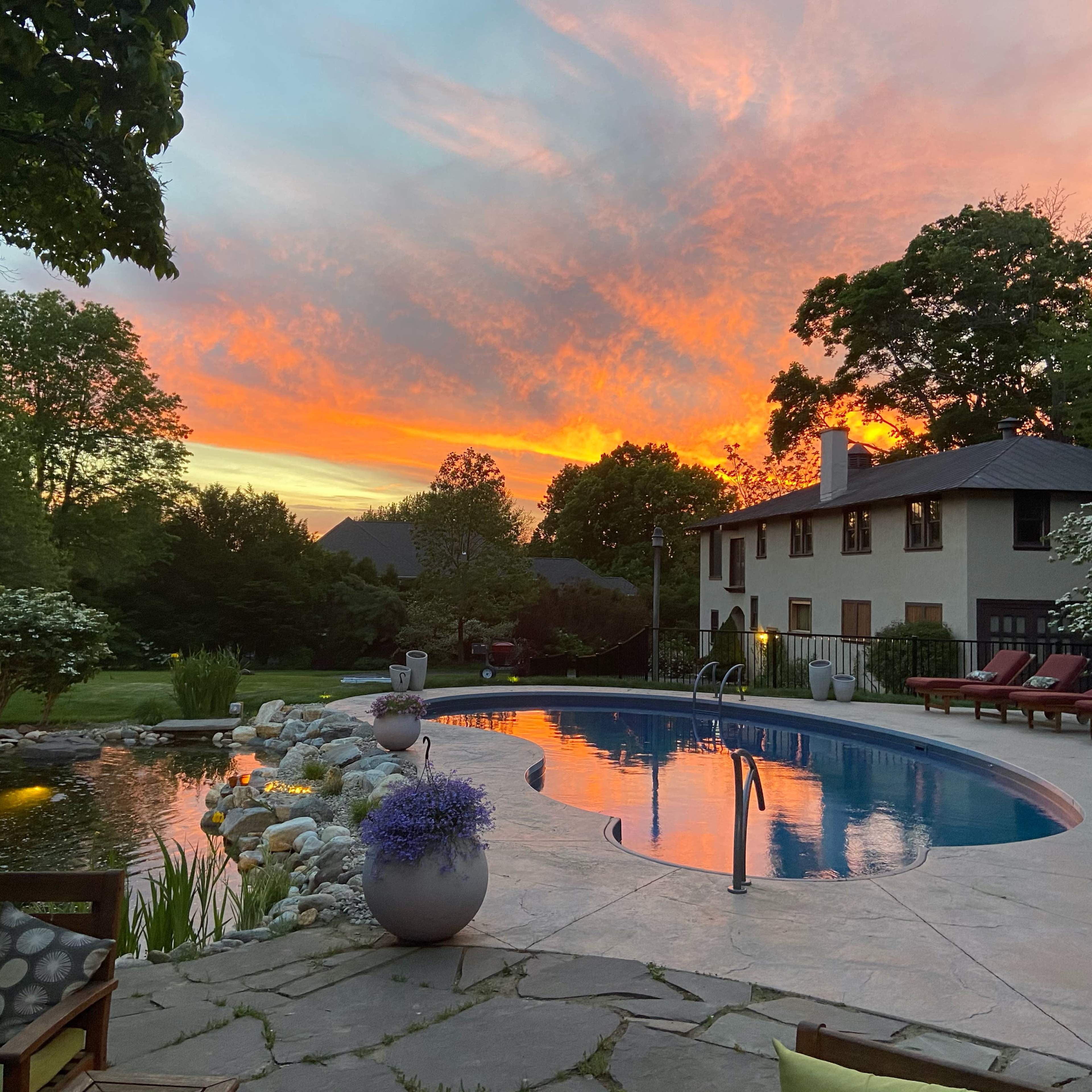 The scene features a swimming pool surrounded by lounge chairs, with a vibrant sunset illuminating the sky in the background.