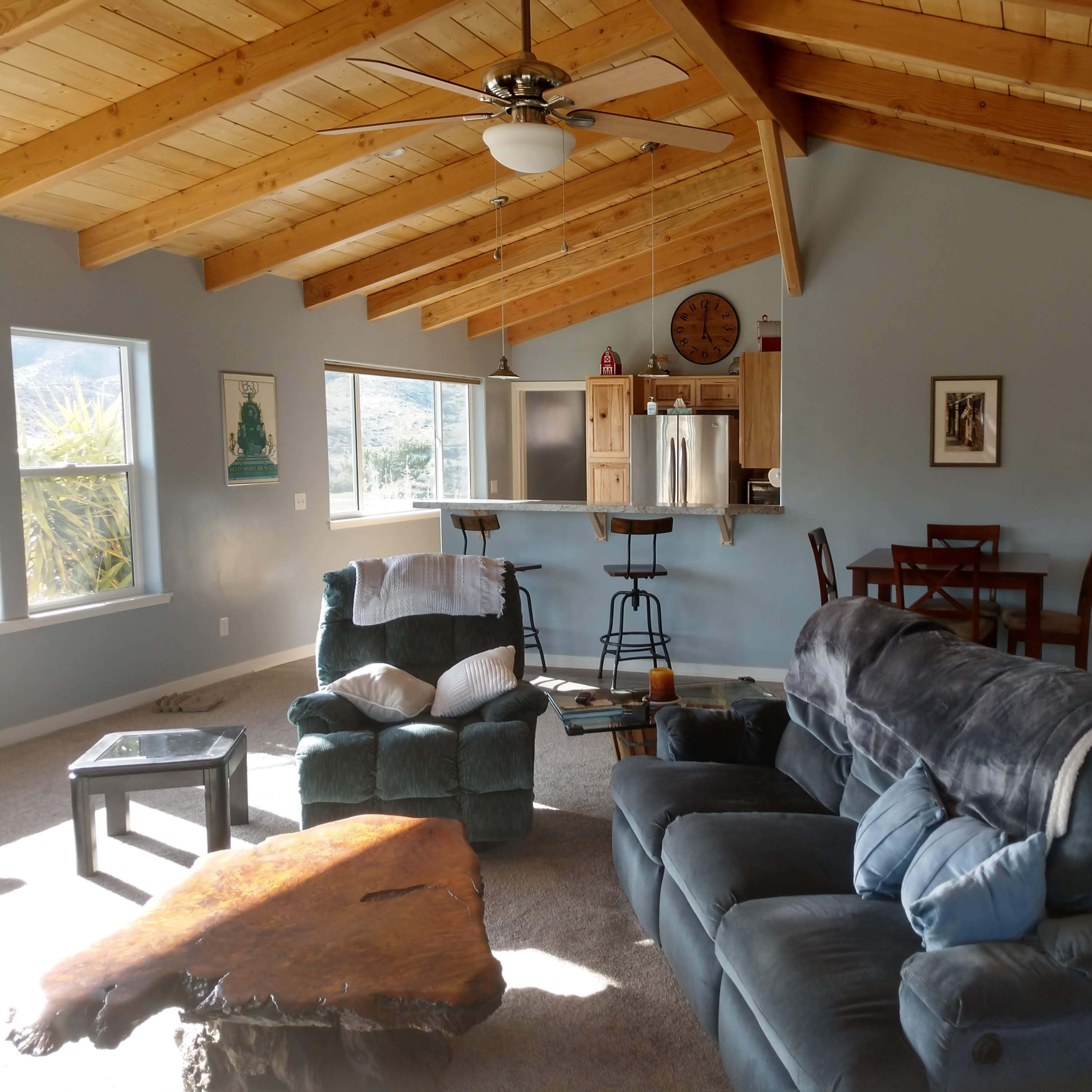 A cozy living room with a vaulted wooden ceiling, featuring a teal loveseat and armchair, a wooden coffee table, and a dining area with a table and chairs in the background.