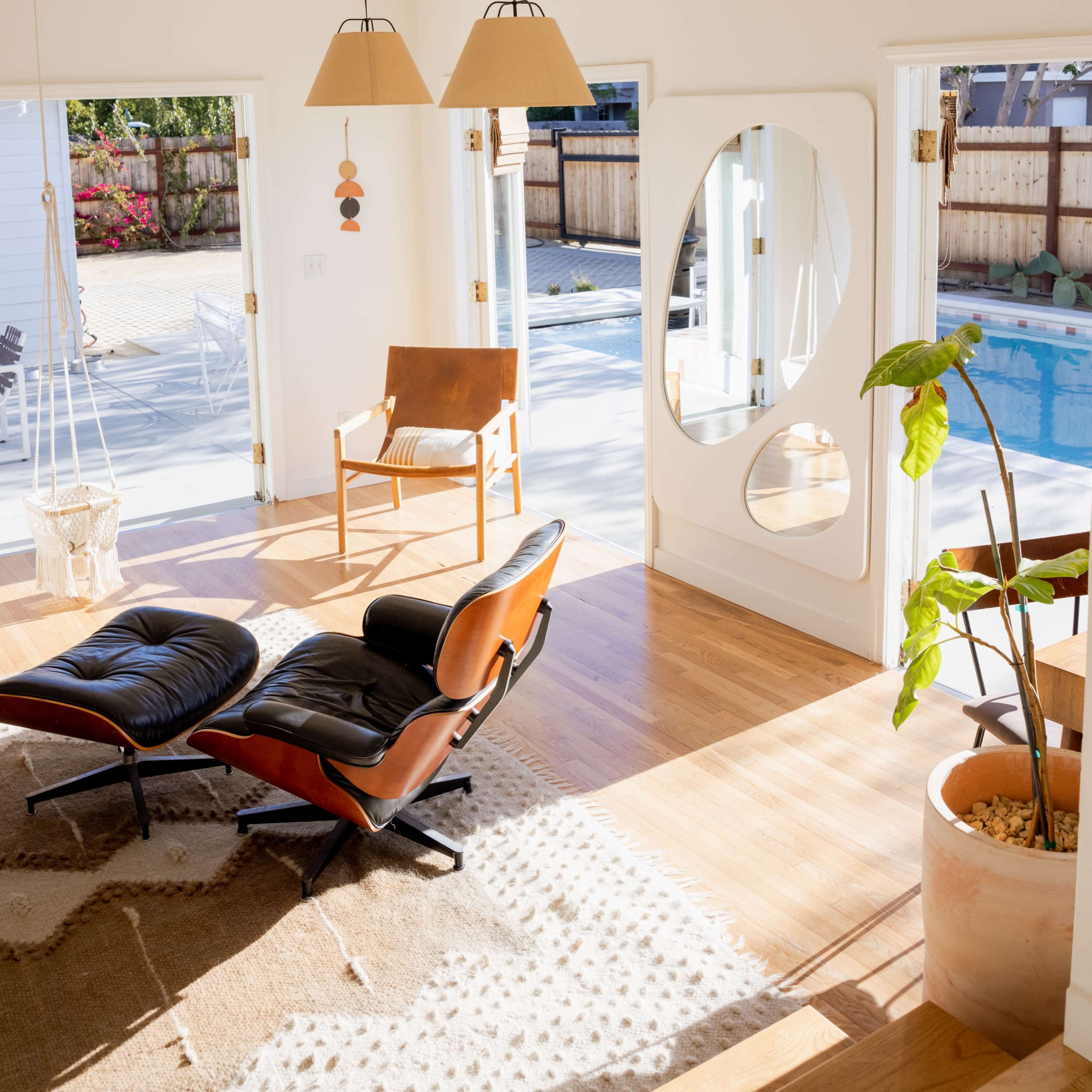 A bright living room features a stylish chair, a large mirror, and open doors leading to a pool area.