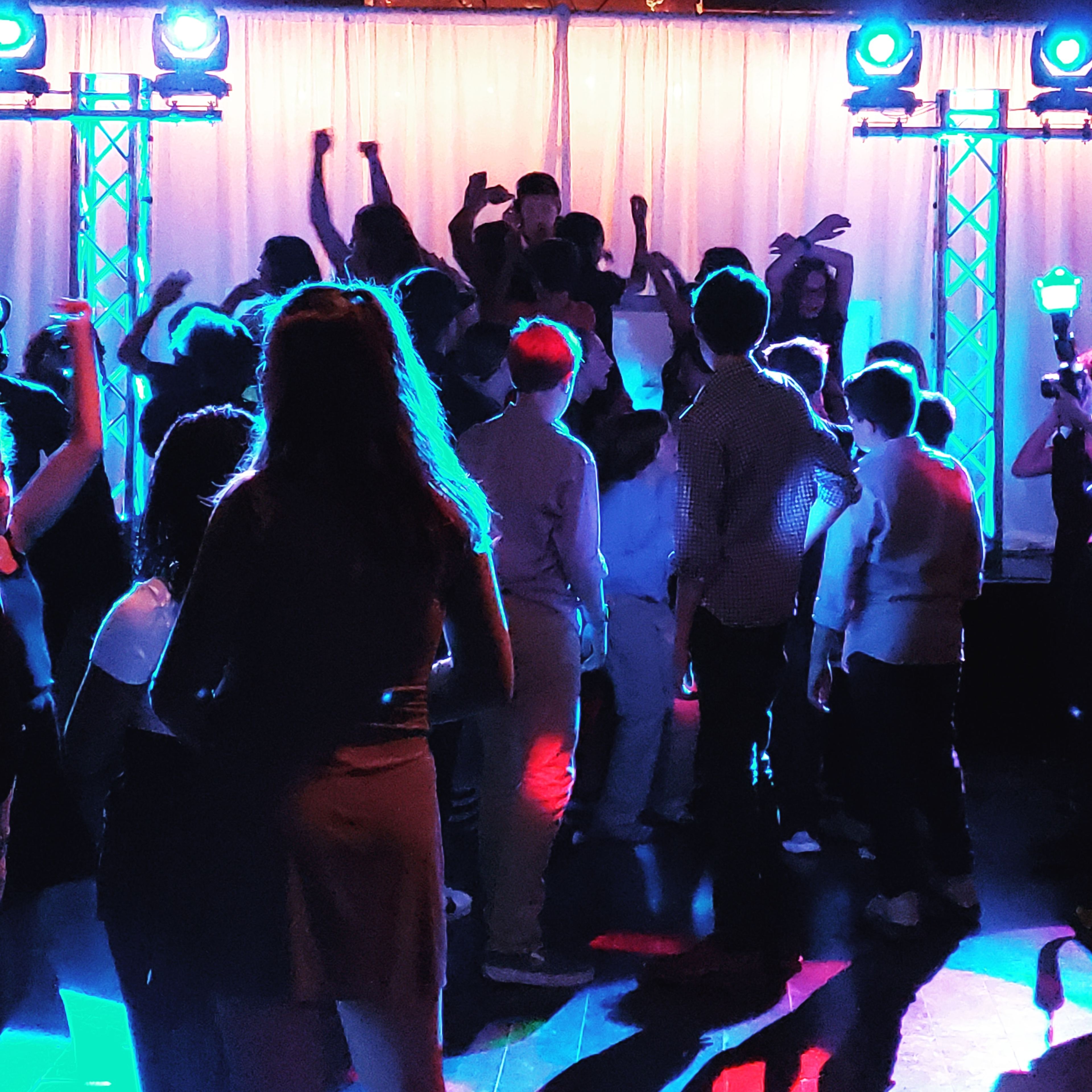 A group of people dance under colorful lights in a venue with draped curtains.