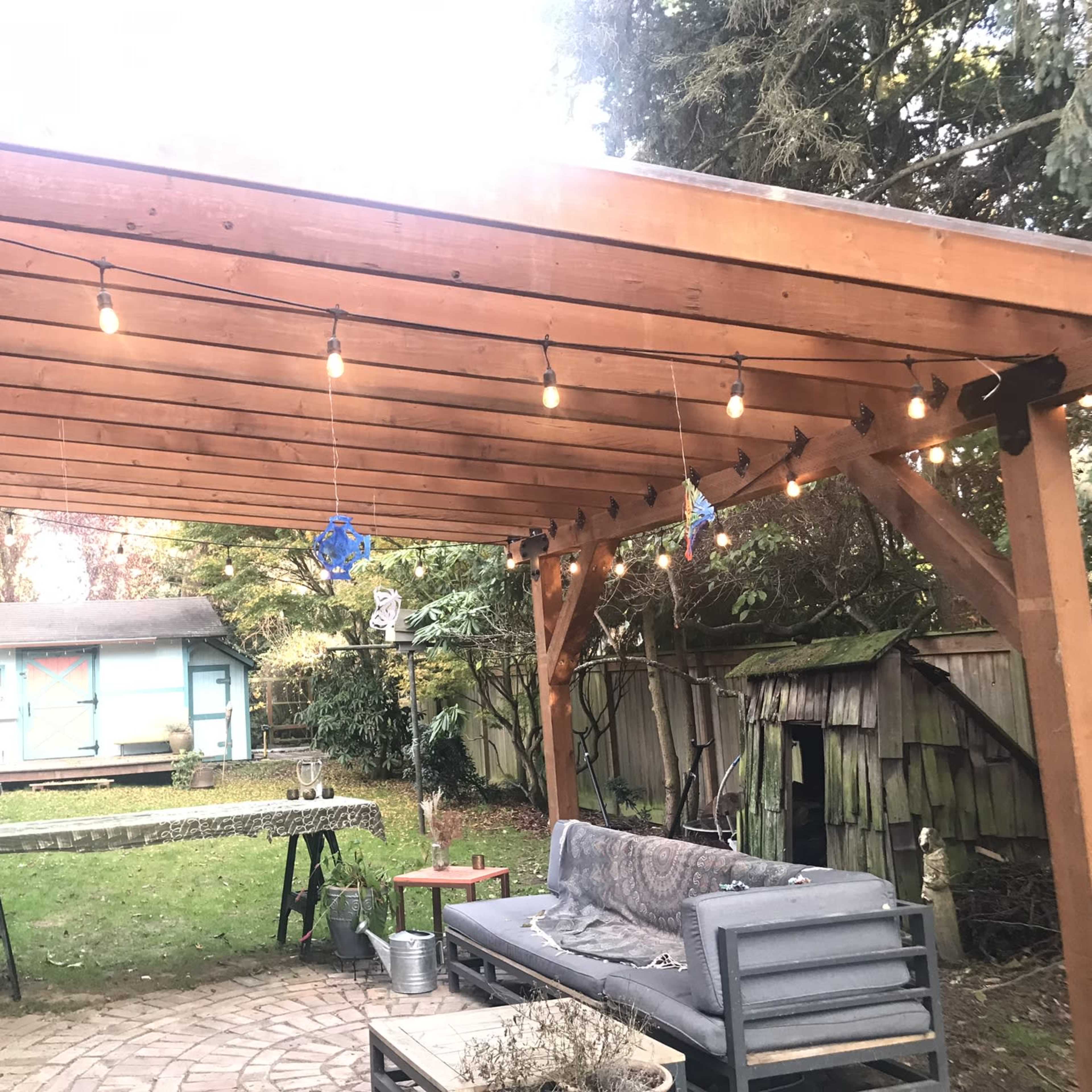 A wooden pergola with hanging lights overlooks a grassy backyard where a covered table and a seating area are arranged.
