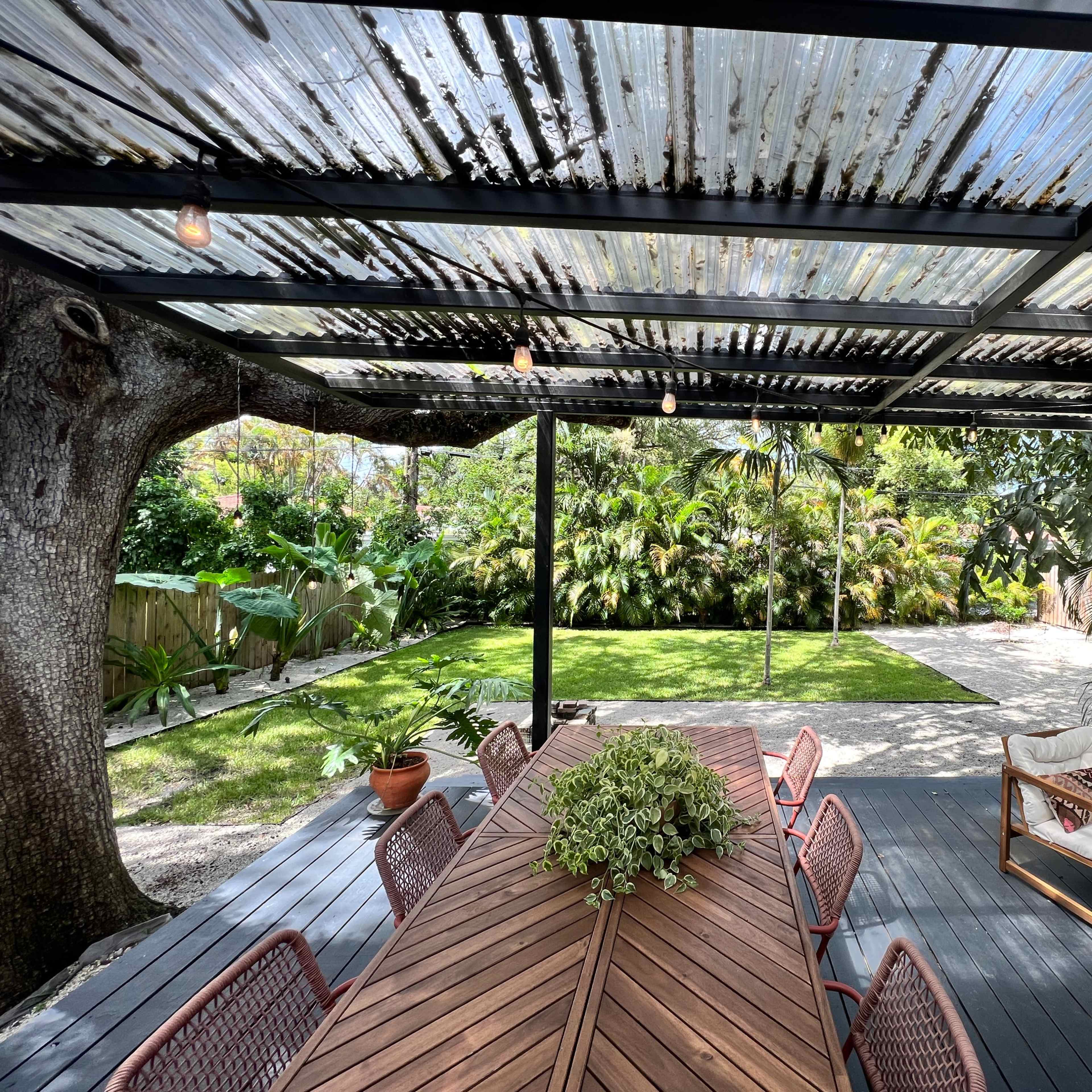 A wooden dining table is set beneath a corrugated metal roof, surrounded by lush greenery in a well-maintained garden.