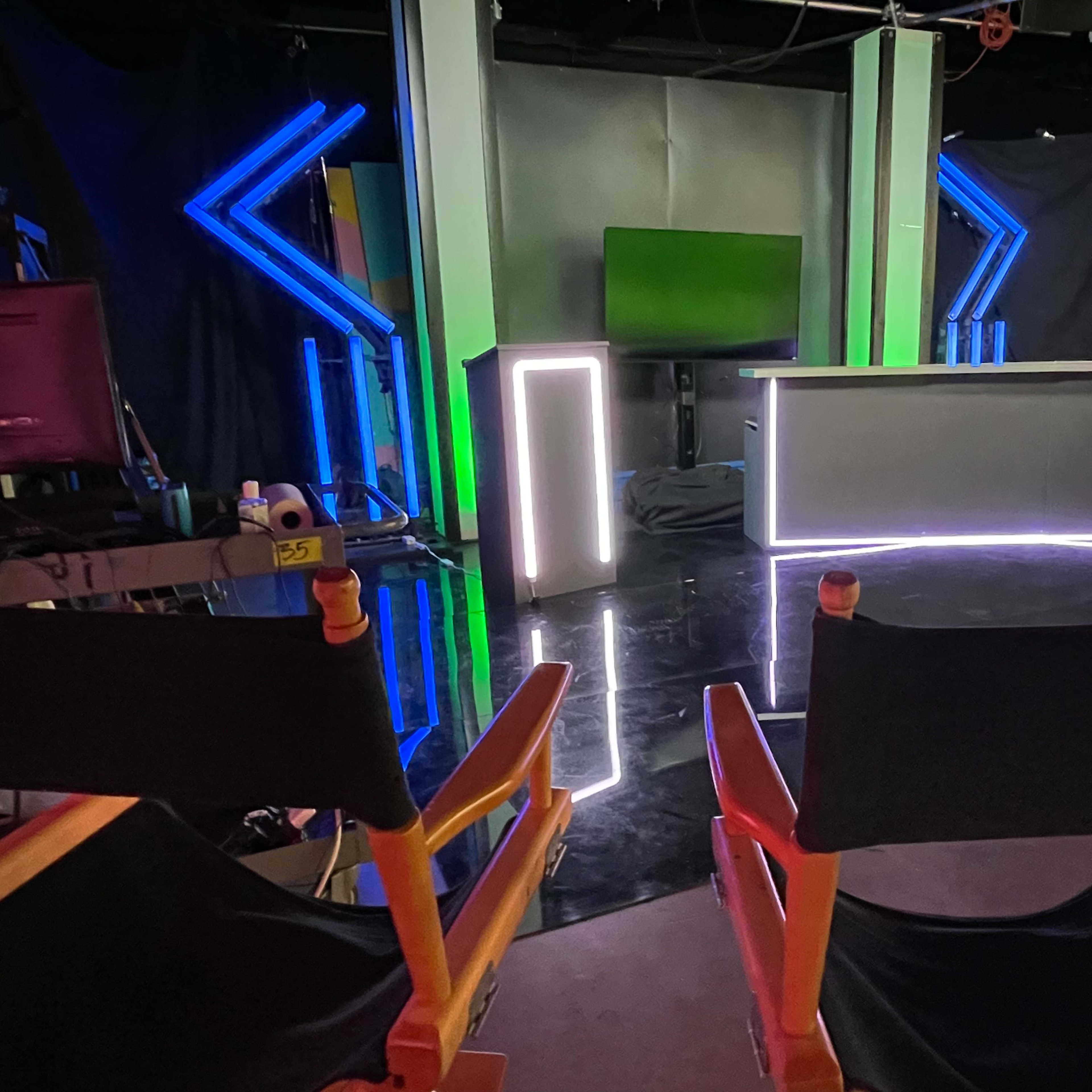 A television studio setup with a lit desk, a backdrop featuring green and blue lights, and two director's chairs in the foreground.