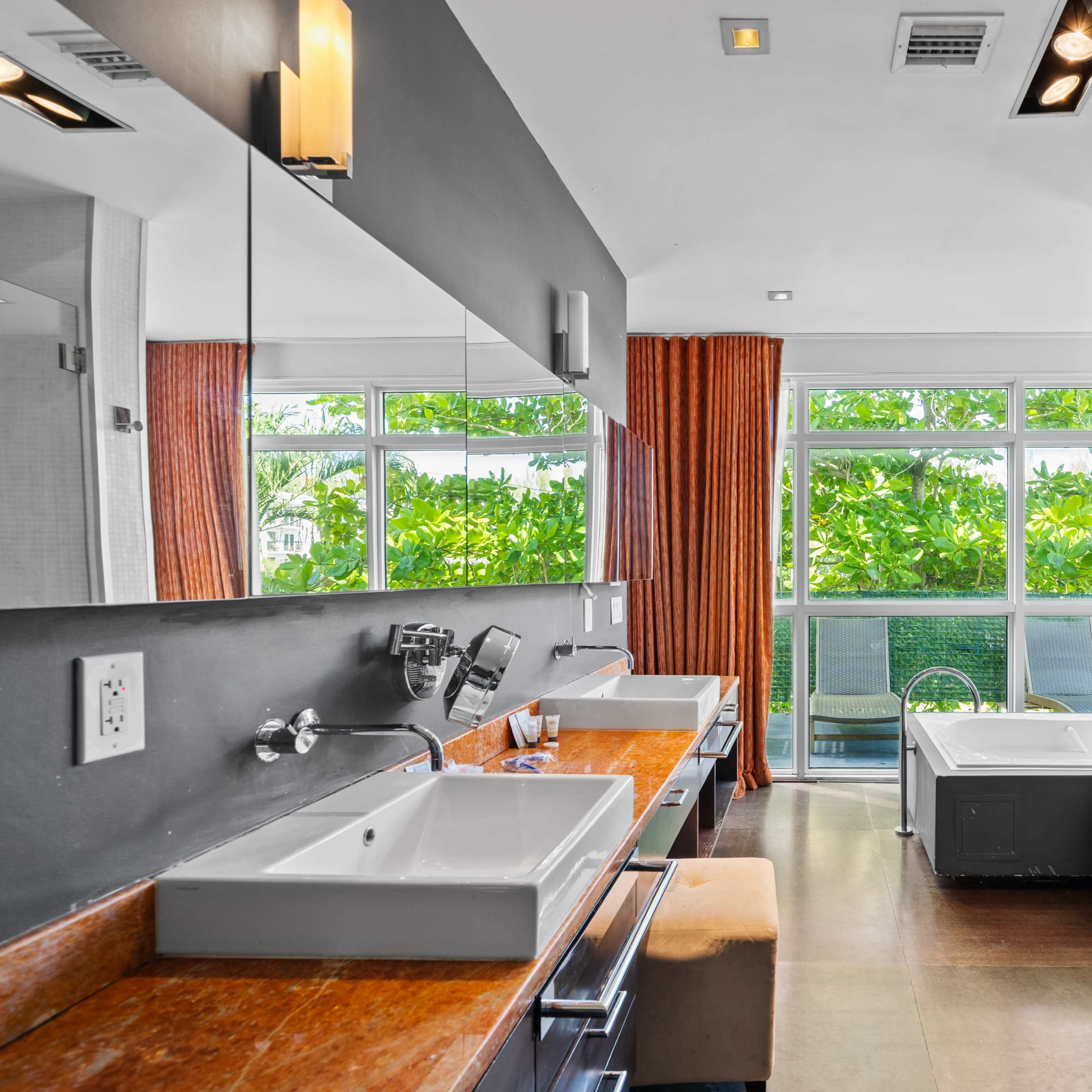 A modern bathroom features dual sinks, a large mirror, and a glass-enclosed shower with a view of greenery outside.