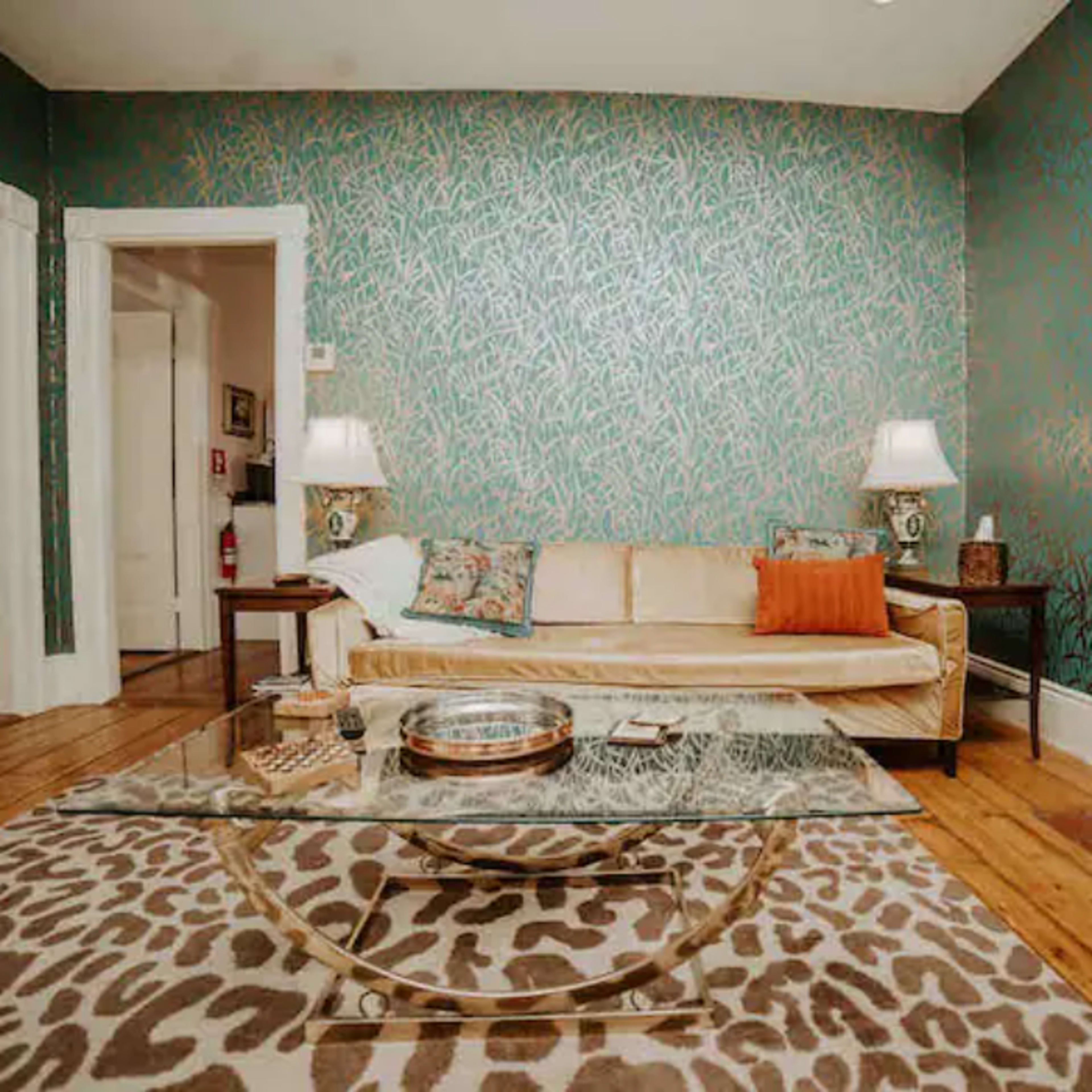 The living room features a patterned wallpaper, a beige sofa with an orange pillow, a glass coffee table, and a fireplace with a brick surround.