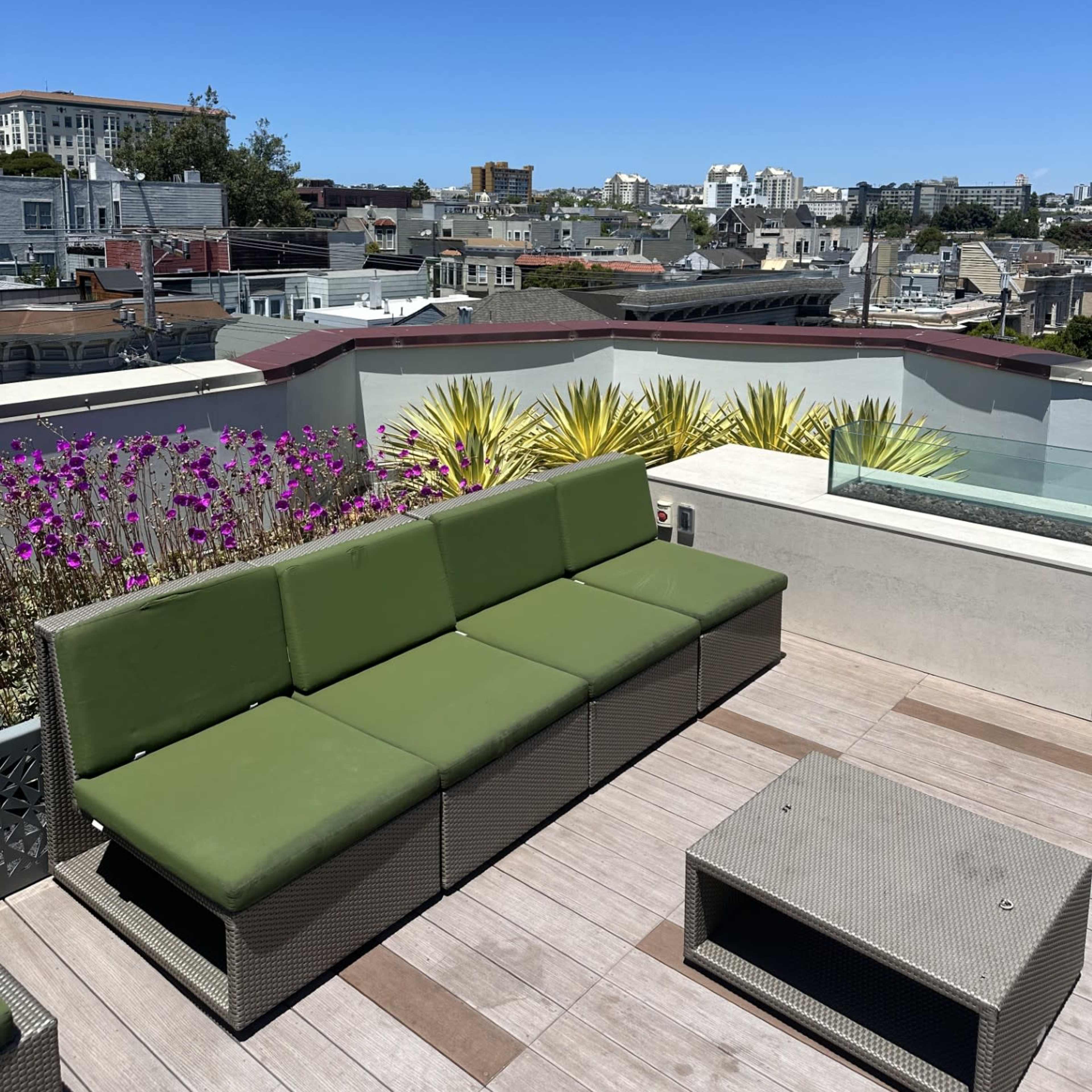 A rooftop lounge area features a green sectional sofa and a small coffee table, overlooking a city skyline under a clear blue sky.