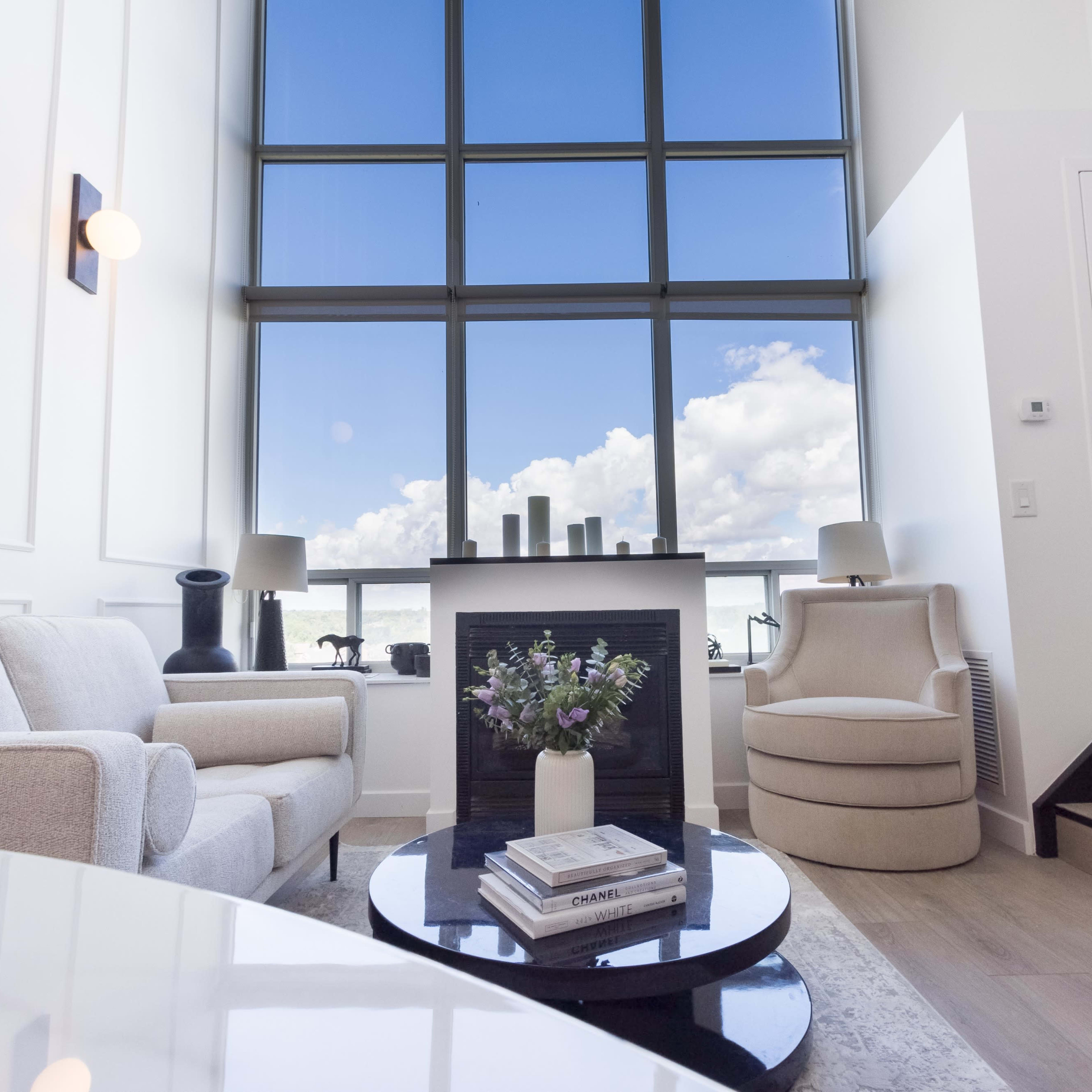 A modern living room features a large window with views of blue skies and clouds, a white sofa, a round chair, and a coffee table with books and a vase of flowers.