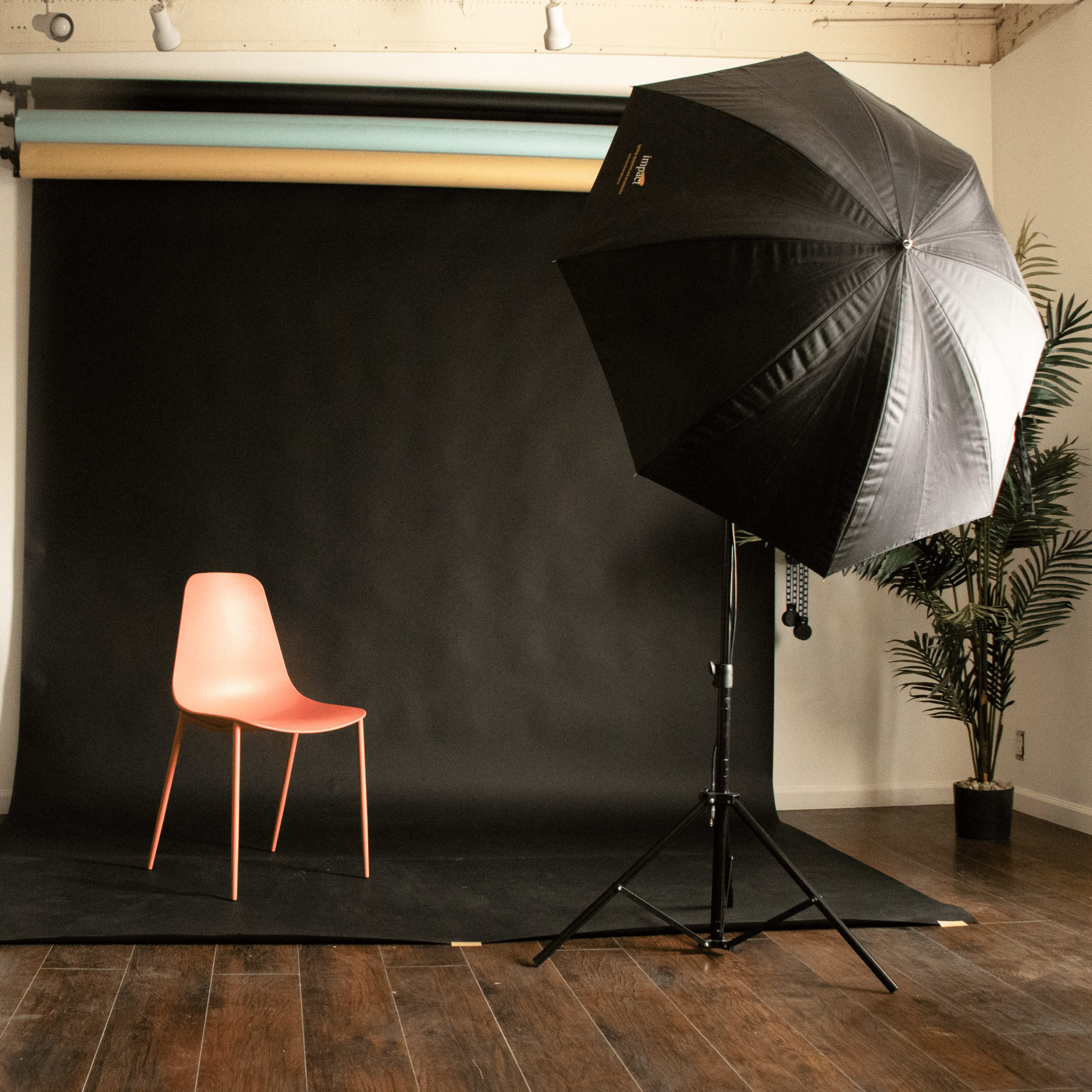 A pink chair is positioned on a dark backdrop in a photography studio, flanked by a gold reflector and a large black umbrella light.