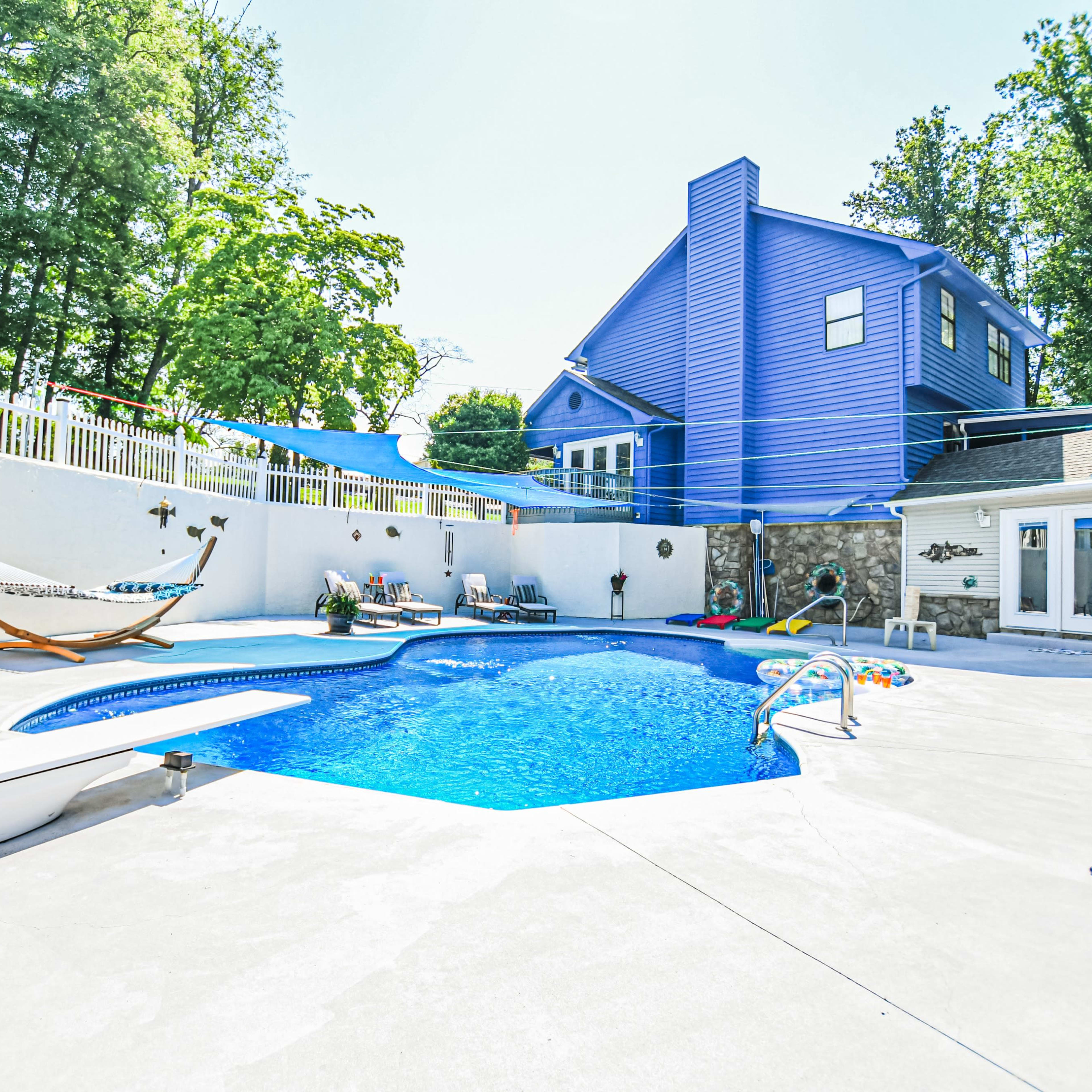 A backyard with a blue swimming pool surrounded by a concrete patio, a hammock, and a house with blue siding in the background.