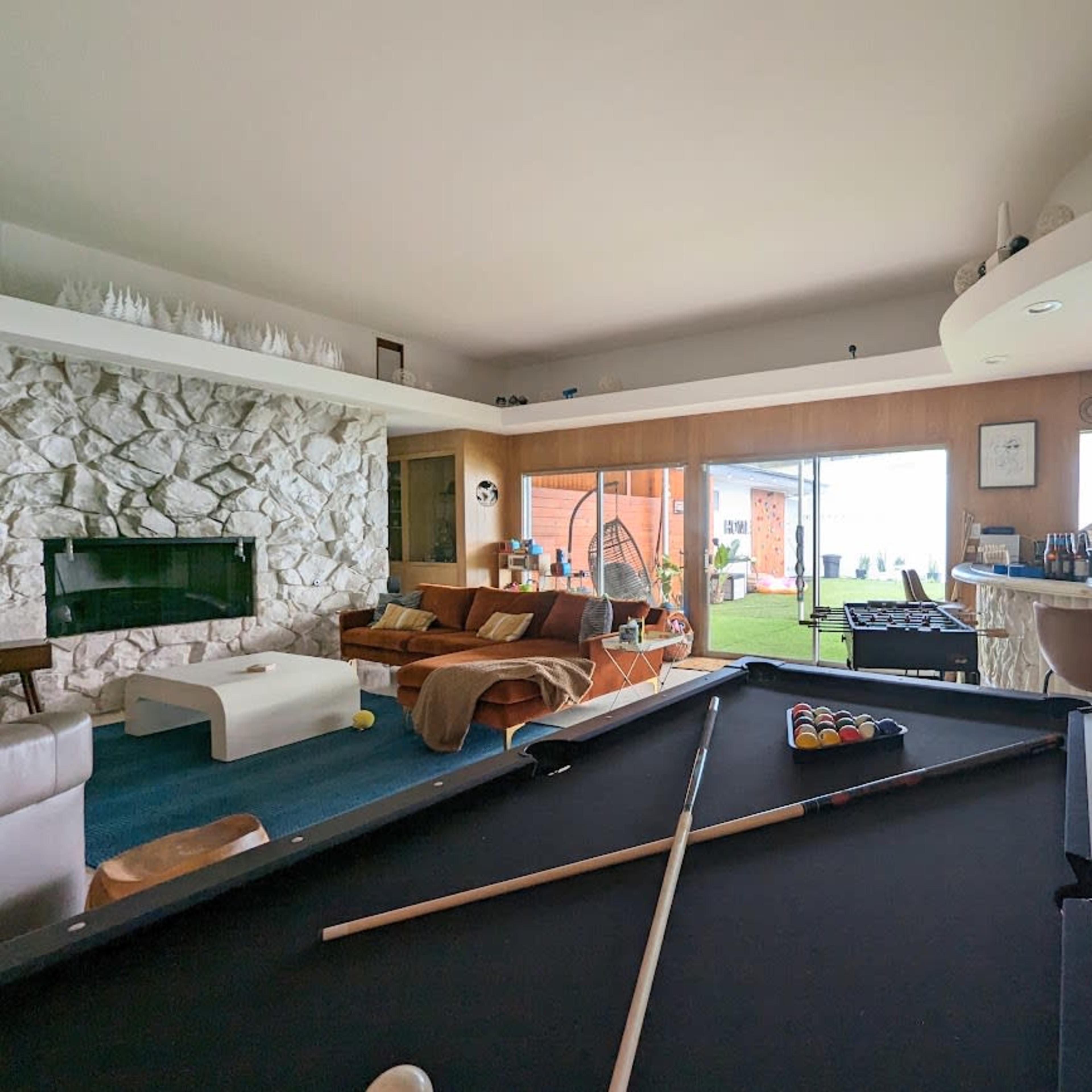 A spacious living area featuring a billiards table in the foreground, a stone fireplace, and a bar with large windows overlooking a grassy outdoor space.