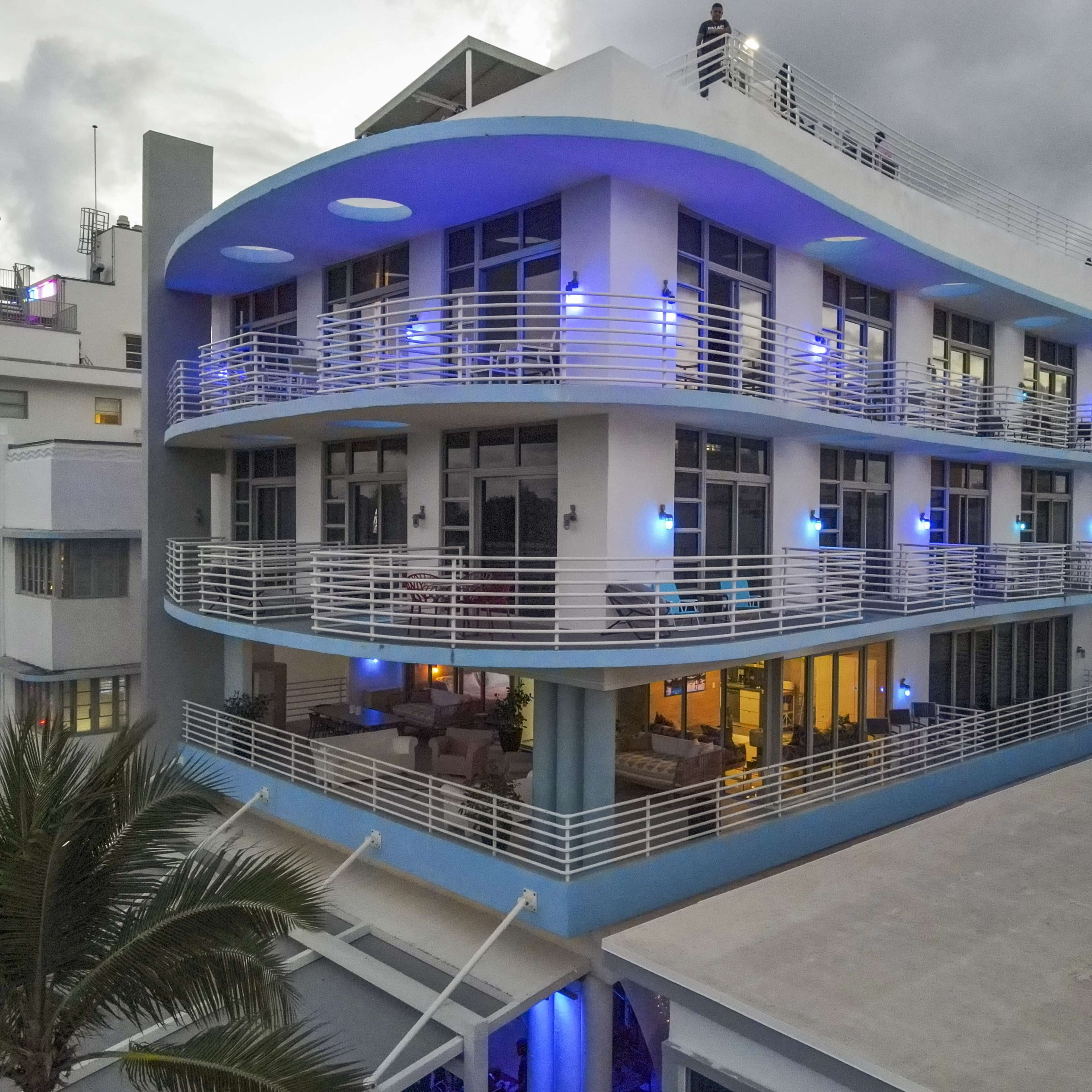 A modern hotel with blue lighting on its balconies and a palm tree in the foreground, set against a cloudy sky.