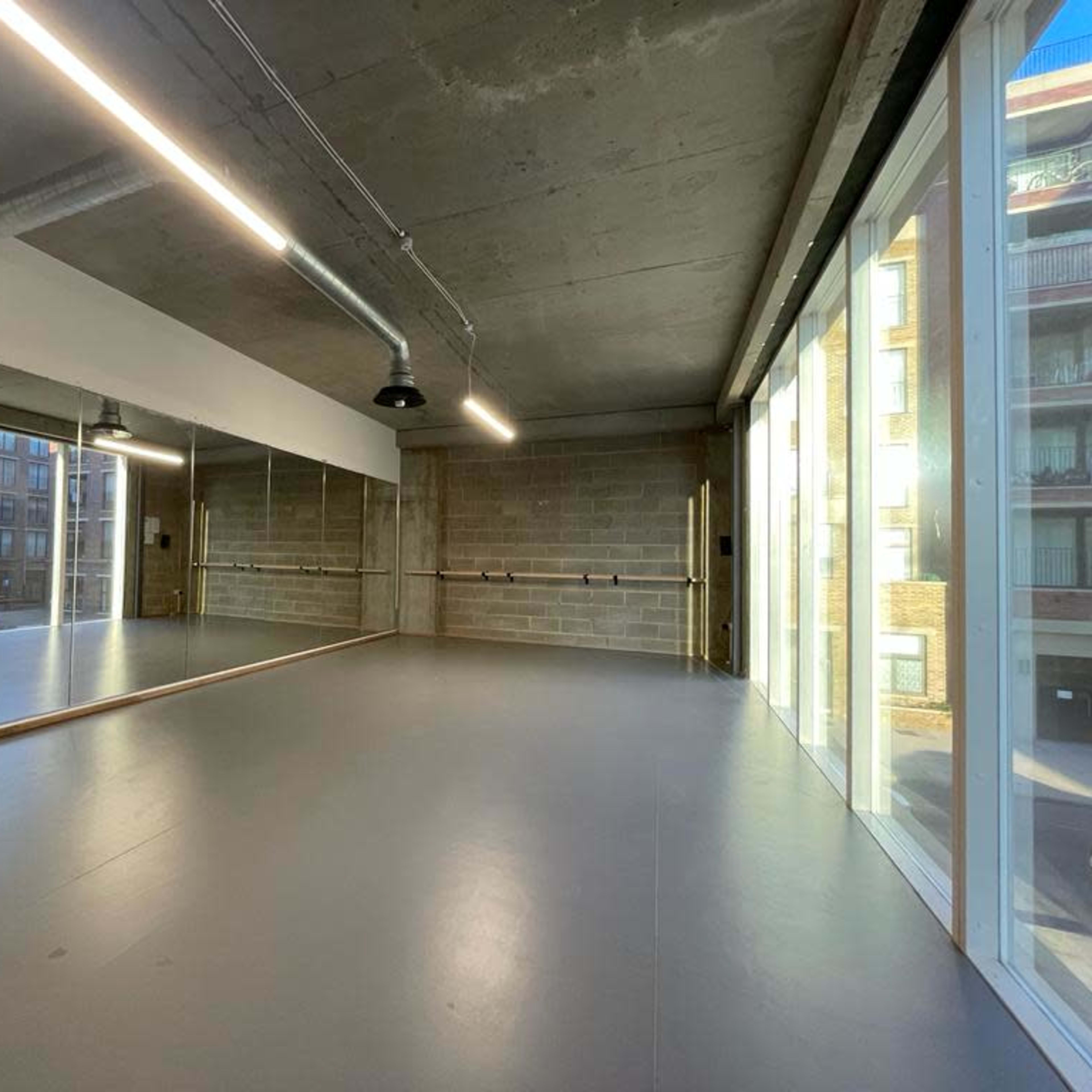 A dance studio features large mirrors on one wall, concrete ceilings, and floor-to-ceiling windows letting in natural light.